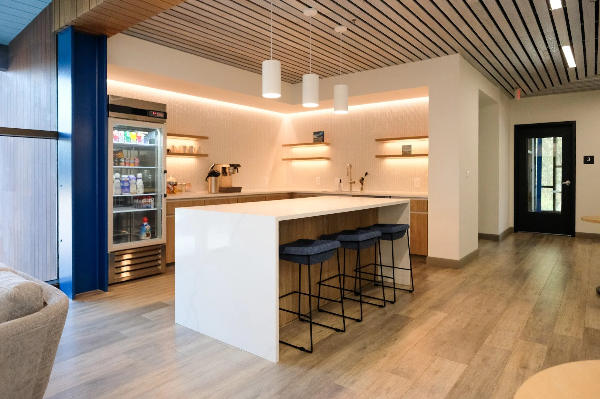 Modern office kitchenette with a white island counter, blue stools, a coffee machine, and a glass-front refrigerator filled with drinks. The room features wooden floors and ceiling panels, and ambient lighting with pendant lights.