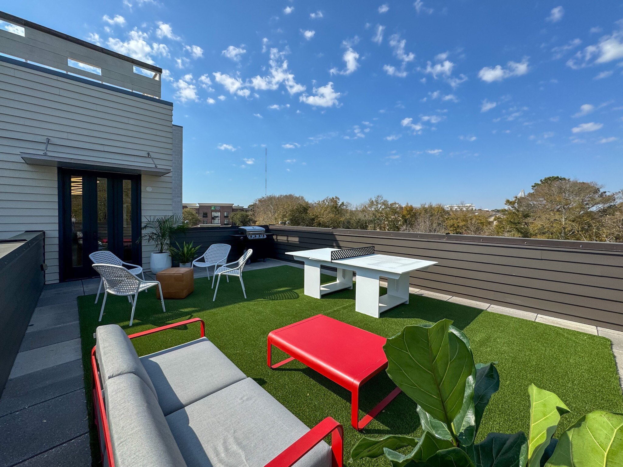 A rooftop terrace with outdoor furniture including a red lounge chair, white armchairs, and a white table, with a grill, potted plants, artificial grass, wooden fencing, and buildings in the background under a blue sky with scattered clouds.