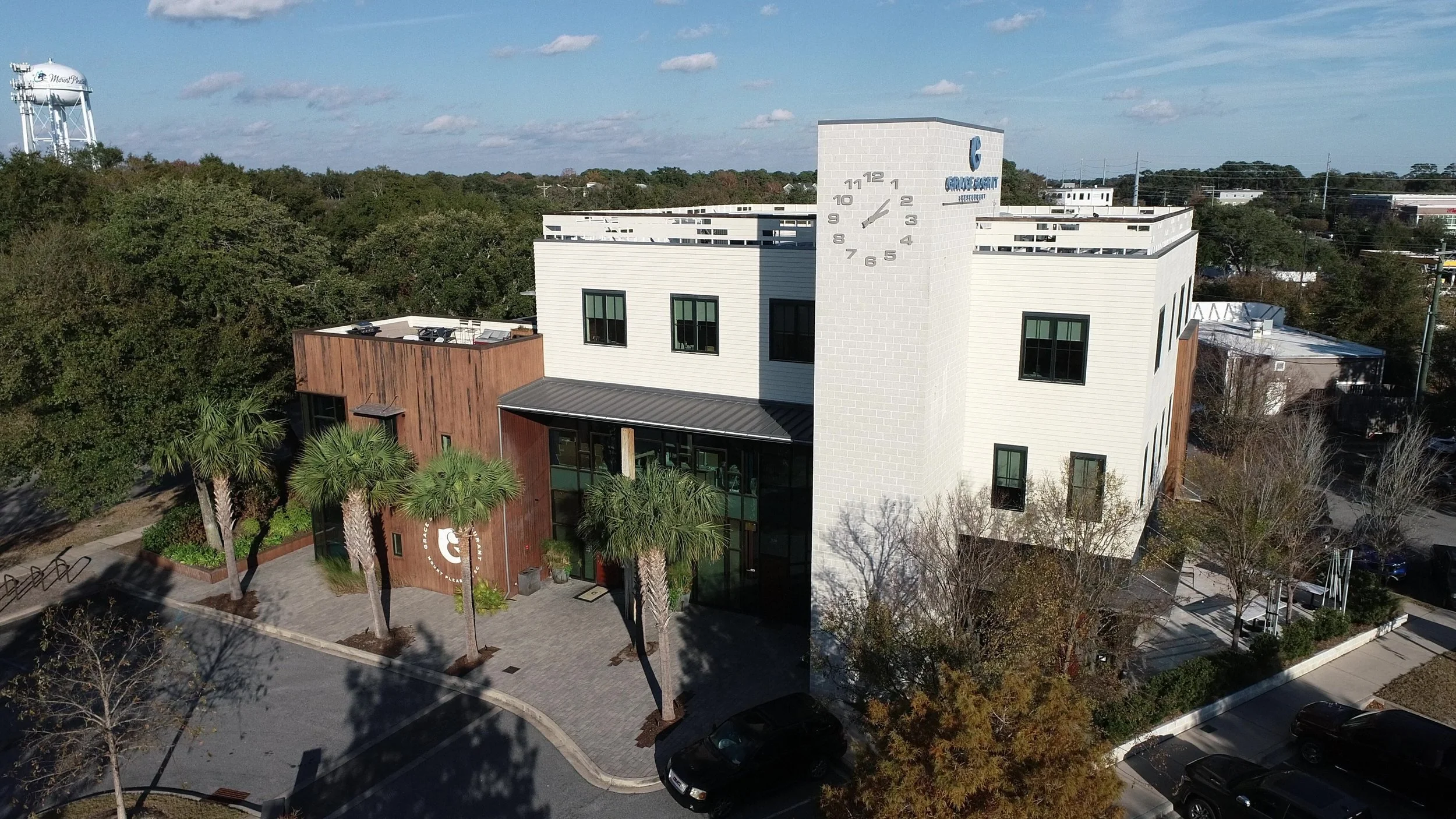 Aerial view of a modern white multi-story building with a clock tower and the logo 'G' for Gracepoint Church, surrounded by trees, parking lots, and a water tower in the distance on a sunny day.