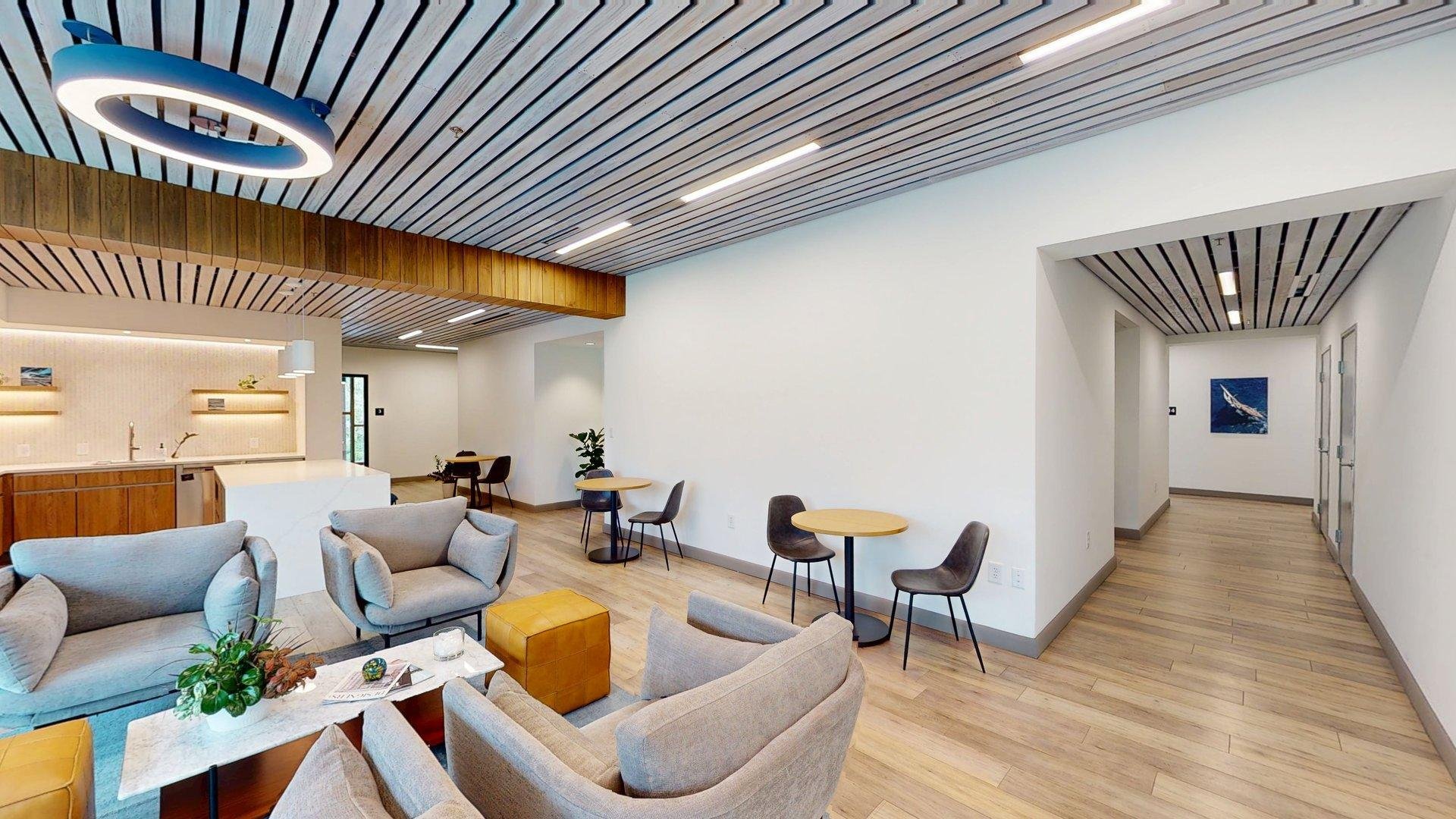 A modern, open-concept living and dining area with wood flooring and white walls. The ceiling features wood and light fixtures. Furniture includes beige armchairs, a sofa, small round tables, and a kitchen with wooden cabinets, a white countertop, and a sink. There are some decorative plants and artwork on the walls.