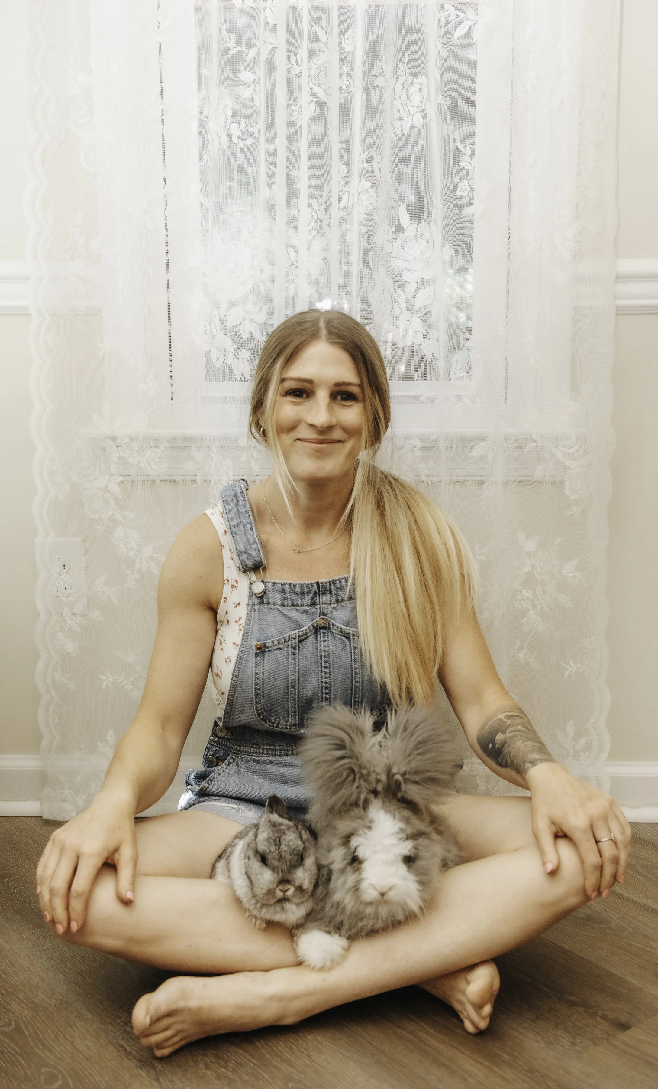 A woman with long blonde hair, wearing denim overalls and a white shirt, sitting cross-legged on a wooden floor. She is smiling and holding three adorable baby bunnies with fluffy fur, sitting on her lap. Behind her, there is a window with lace curtains and white walls.
