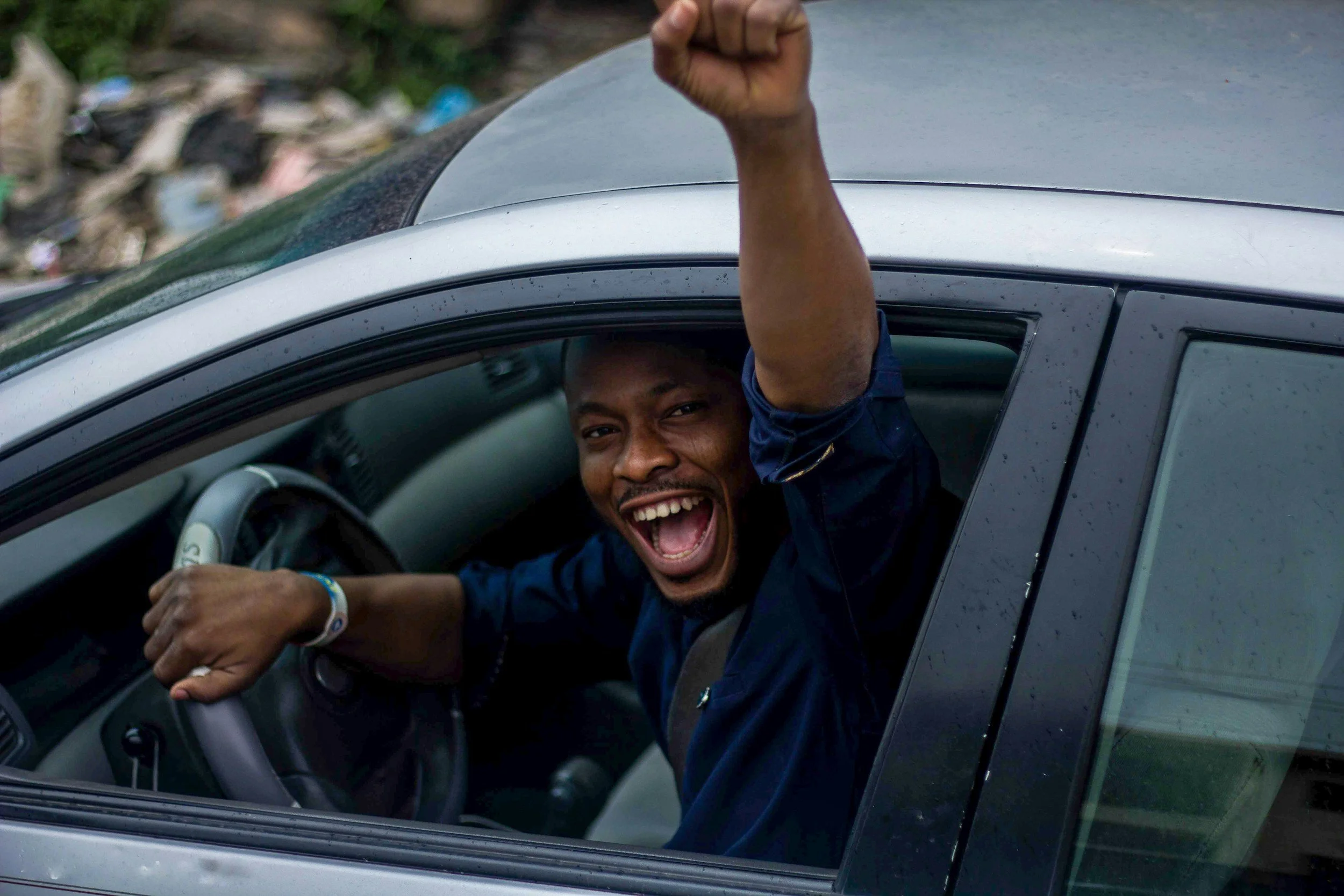 A joyful man sitting in a car, smiling with his mouth open, raising his fist out of the window.