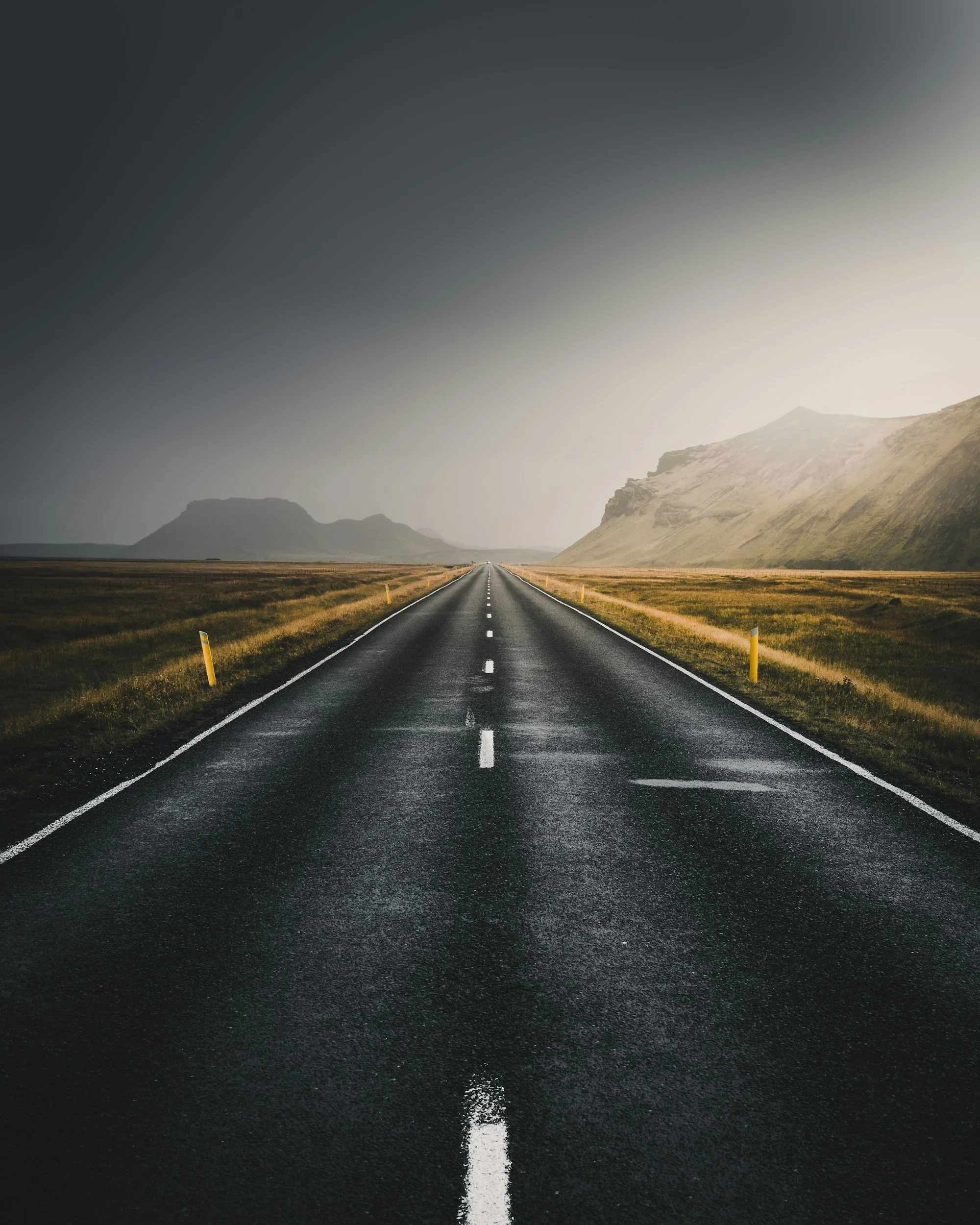 A straight open road stretching toward distant mountains under a cloudy sky with a gradient from dark to light.