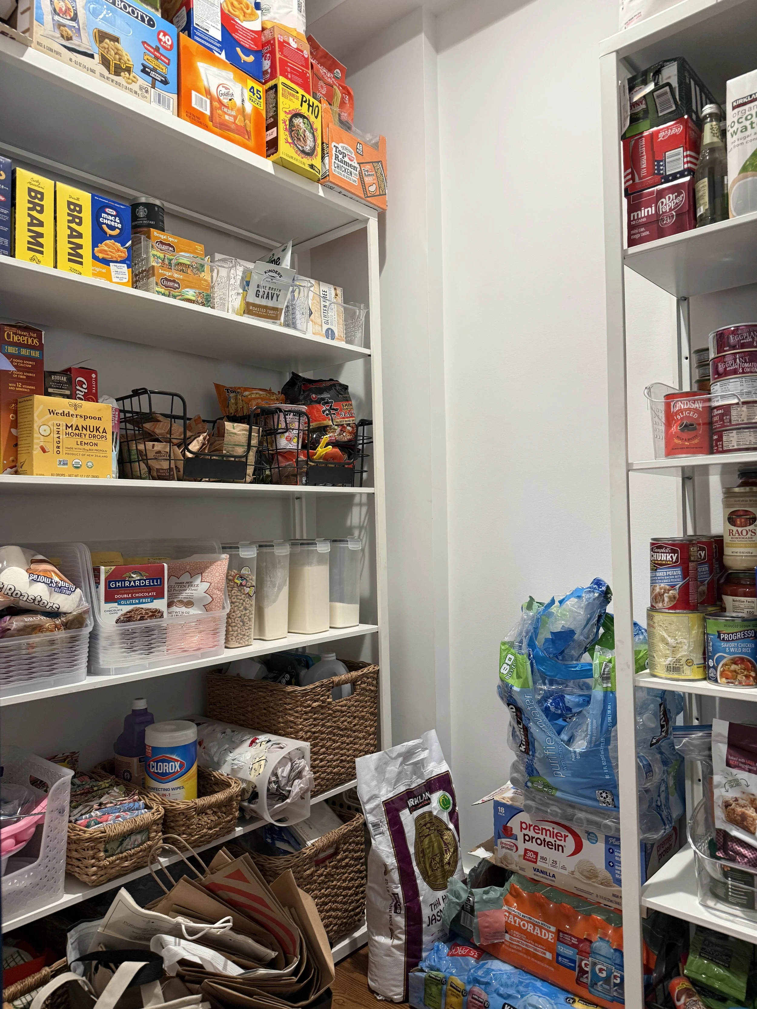 Pantry with white shelves filled with snacks, canned goods, and household items. Baskets and bags are on the floor and shelves.