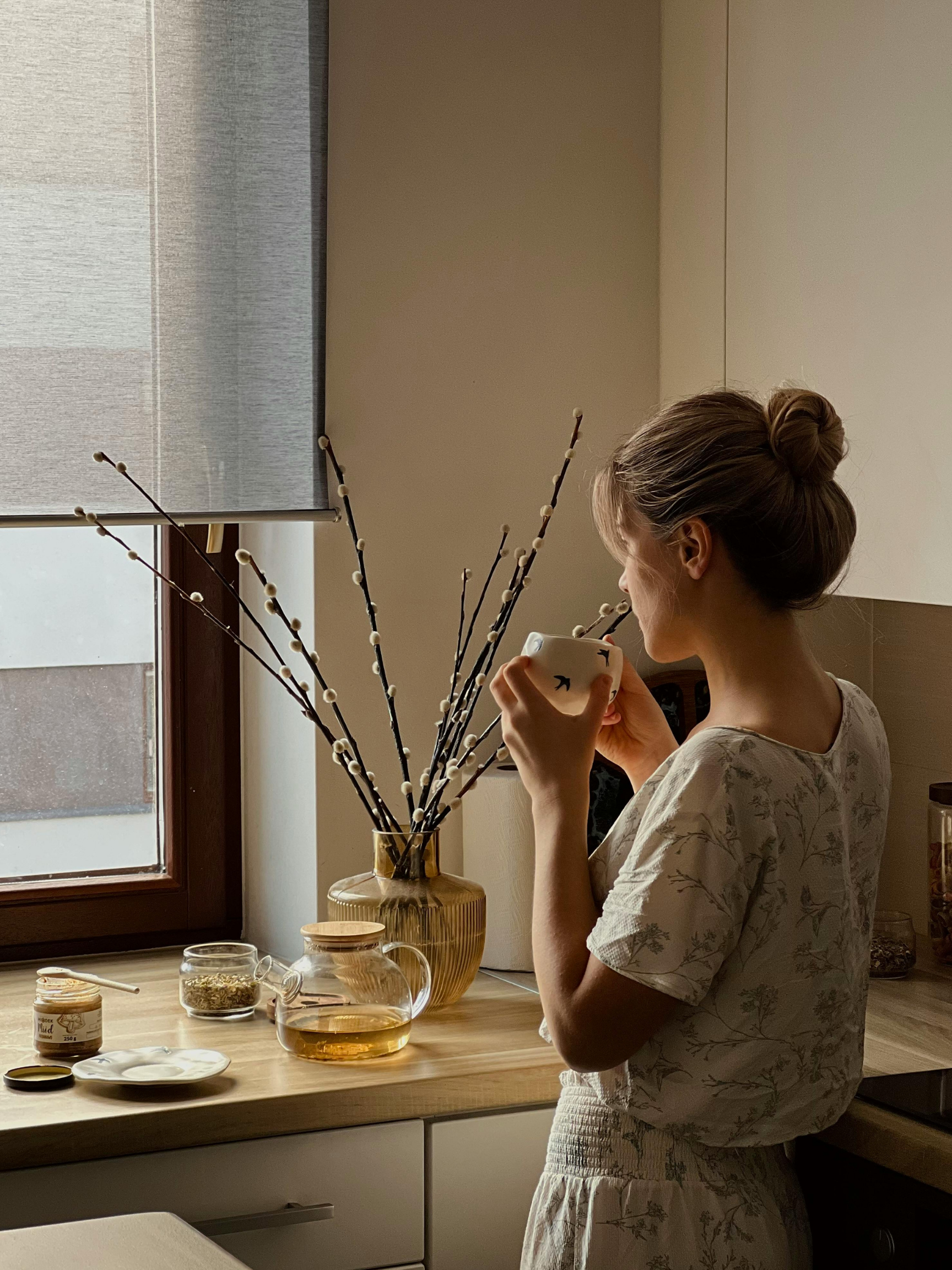 Woman with bun in a kitchen, holding a mug near a window with a light gray roller shade. On the counter are jars, a glass pitcher, and a vase with branches.
