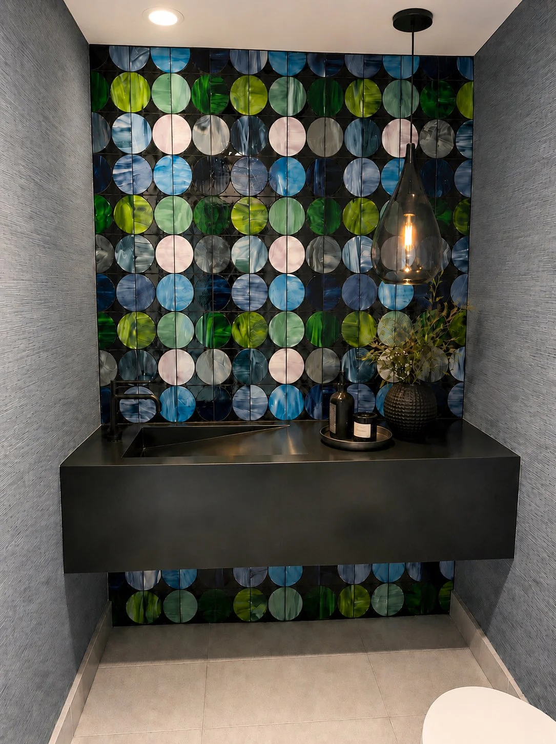 A modern bathroom with dark textured walls, a black floating sink, a textured black vase with green plant, a pendant light, and a colorful circular tile accent wall in shades of green, blue, pink, and gray.