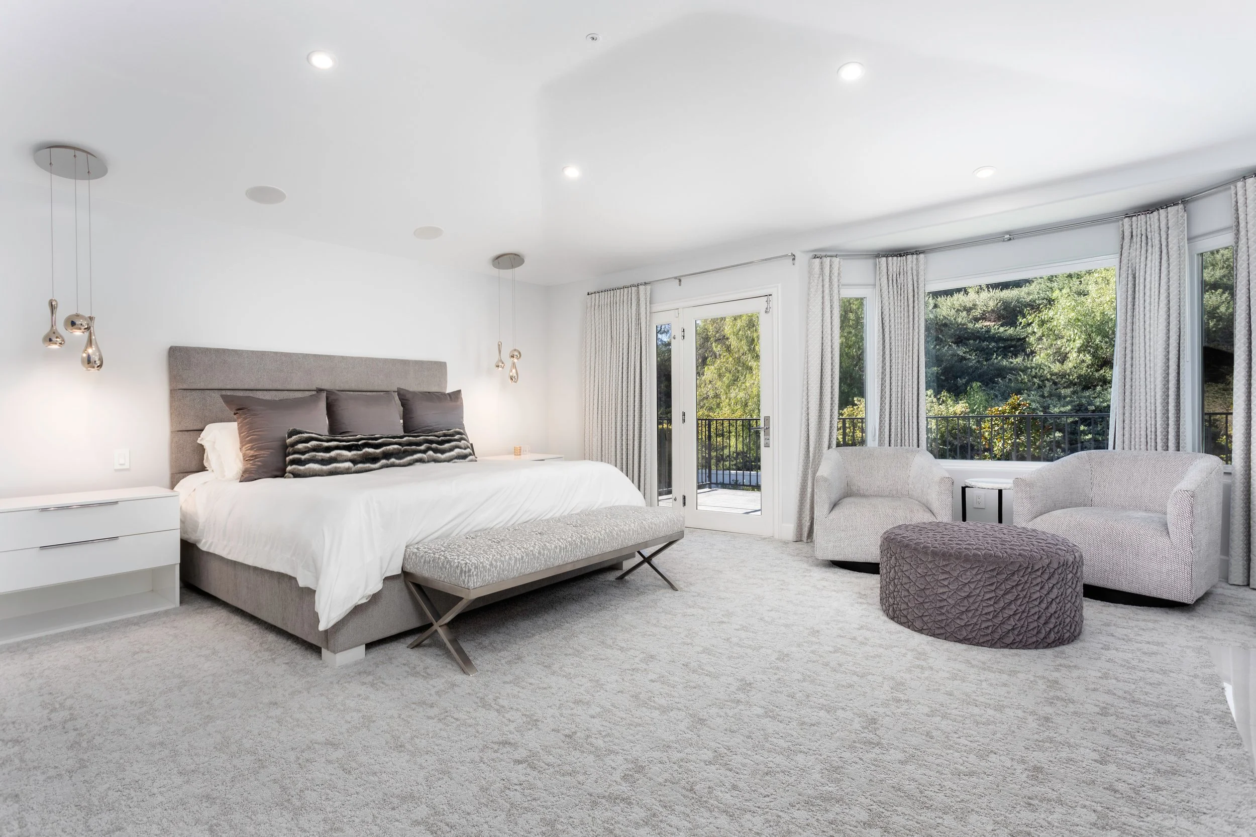 A modern bedroom with a large bed, bedside tables, ceiling lights, and large windows with curtains overlooking greenery.