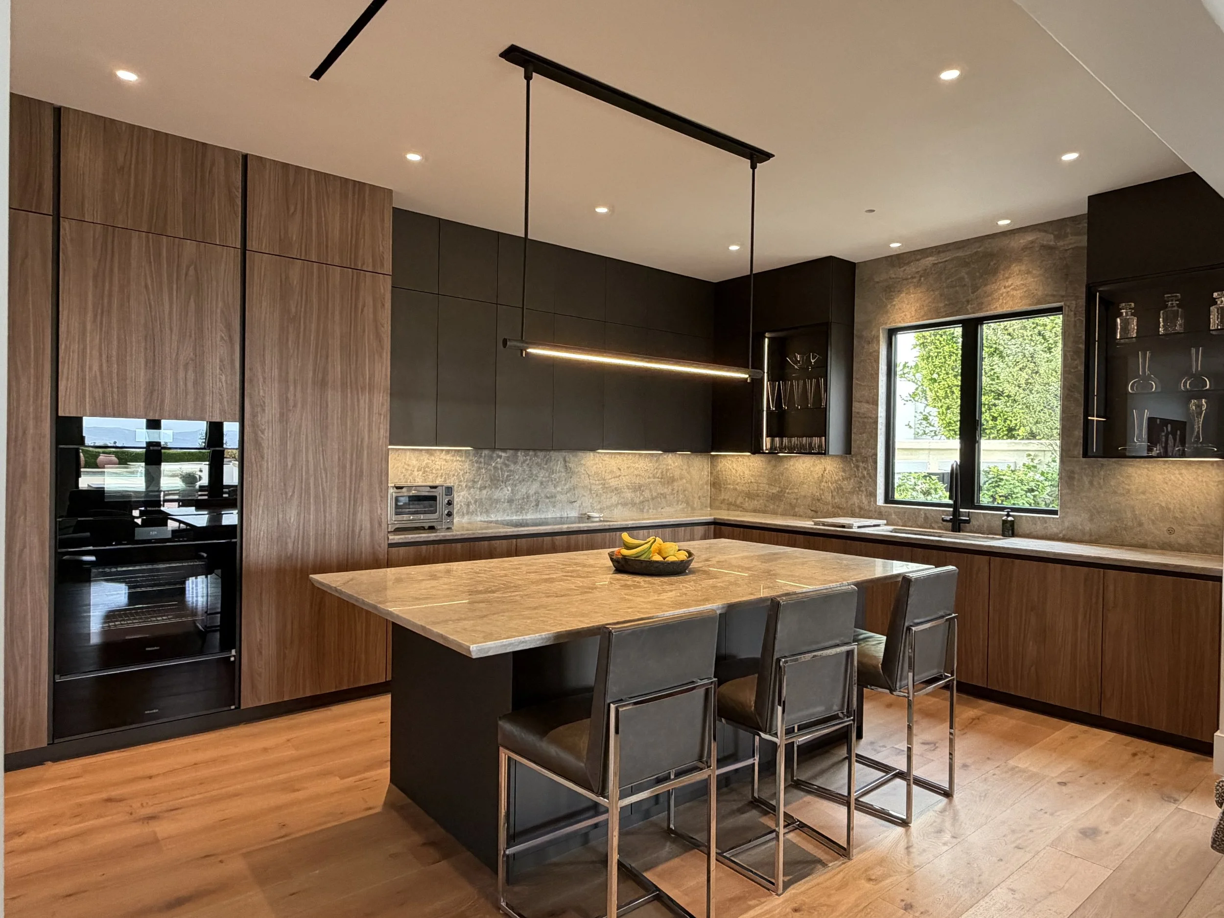 Modern kitchen with wooden cabinets, black and gray accents, a kitchen island with seating, and a bowl of bananas on the island. Window showing green trees outside.