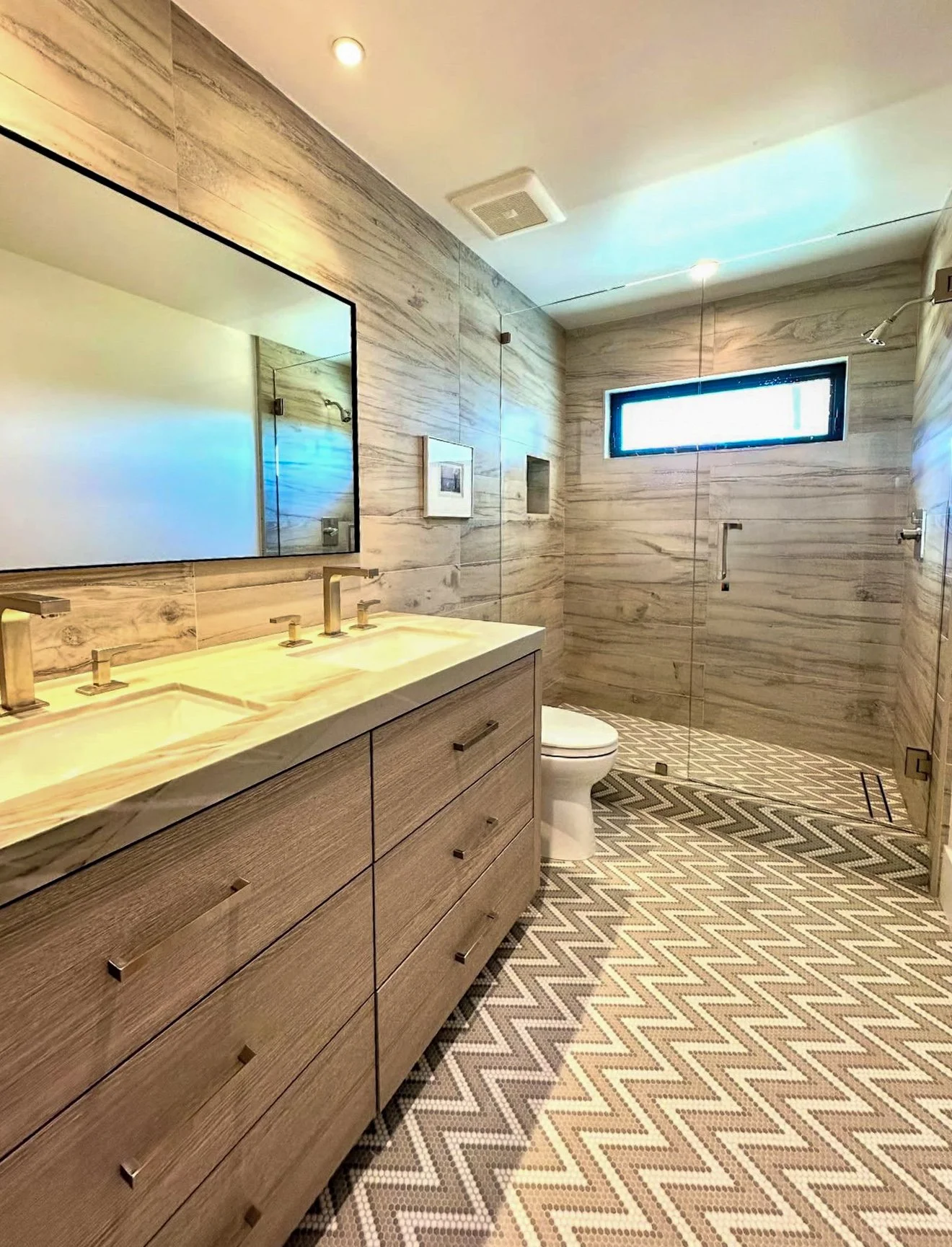 Modern bathroom with wooden vanity, double sink, large mirror, toilet, walk-in shower with glass door, window, and geometric patterned tile floor.