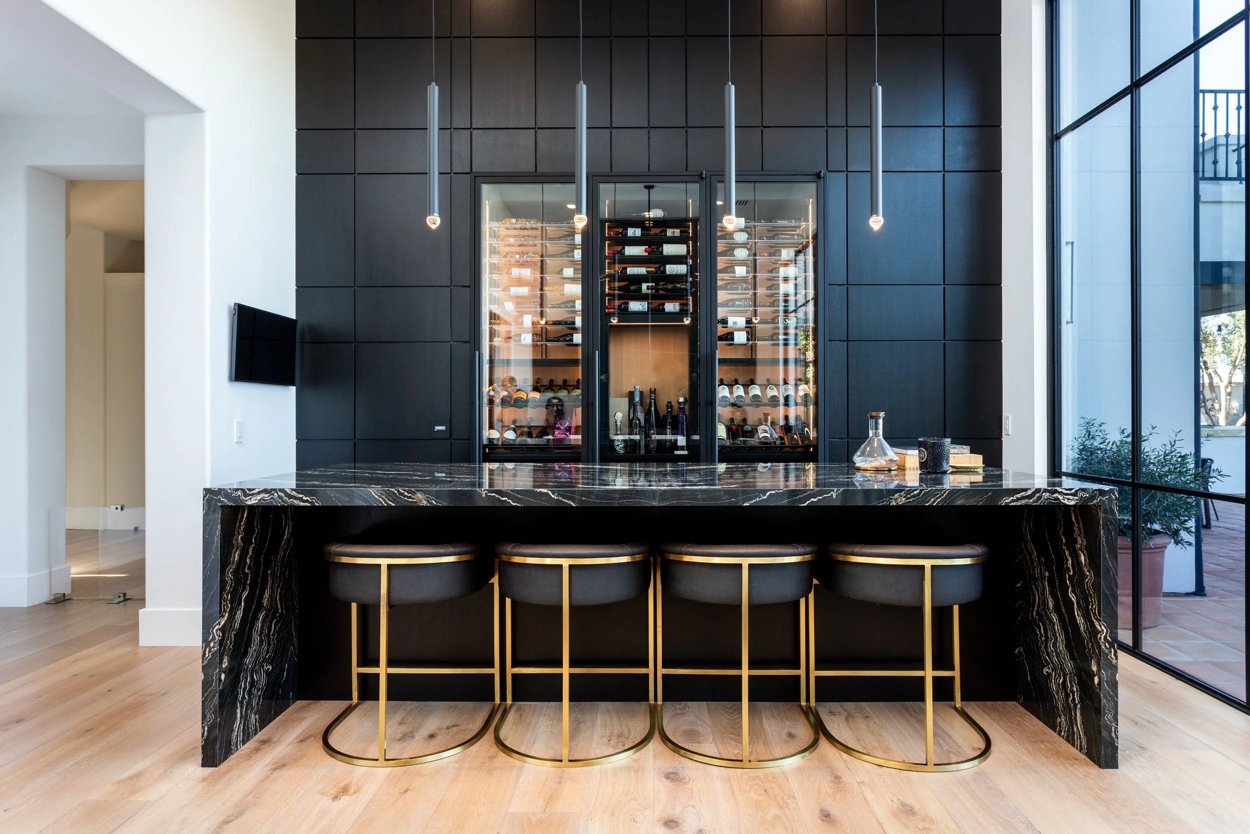 Modern bar area with a black marble countertop, four black and gold bar stools, and a black wall with a glass wine storage cabinet. Large windows on the right let in natural light.