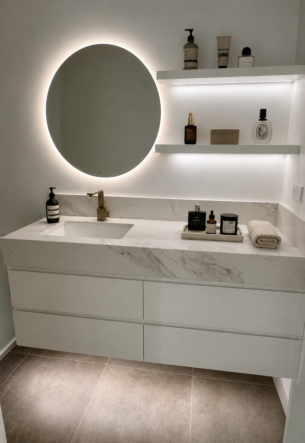 Modern bathroom vanity with a white marble countertop, a brass faucet, a circular backlit mirror, and shelves with skincare products and perfumes.