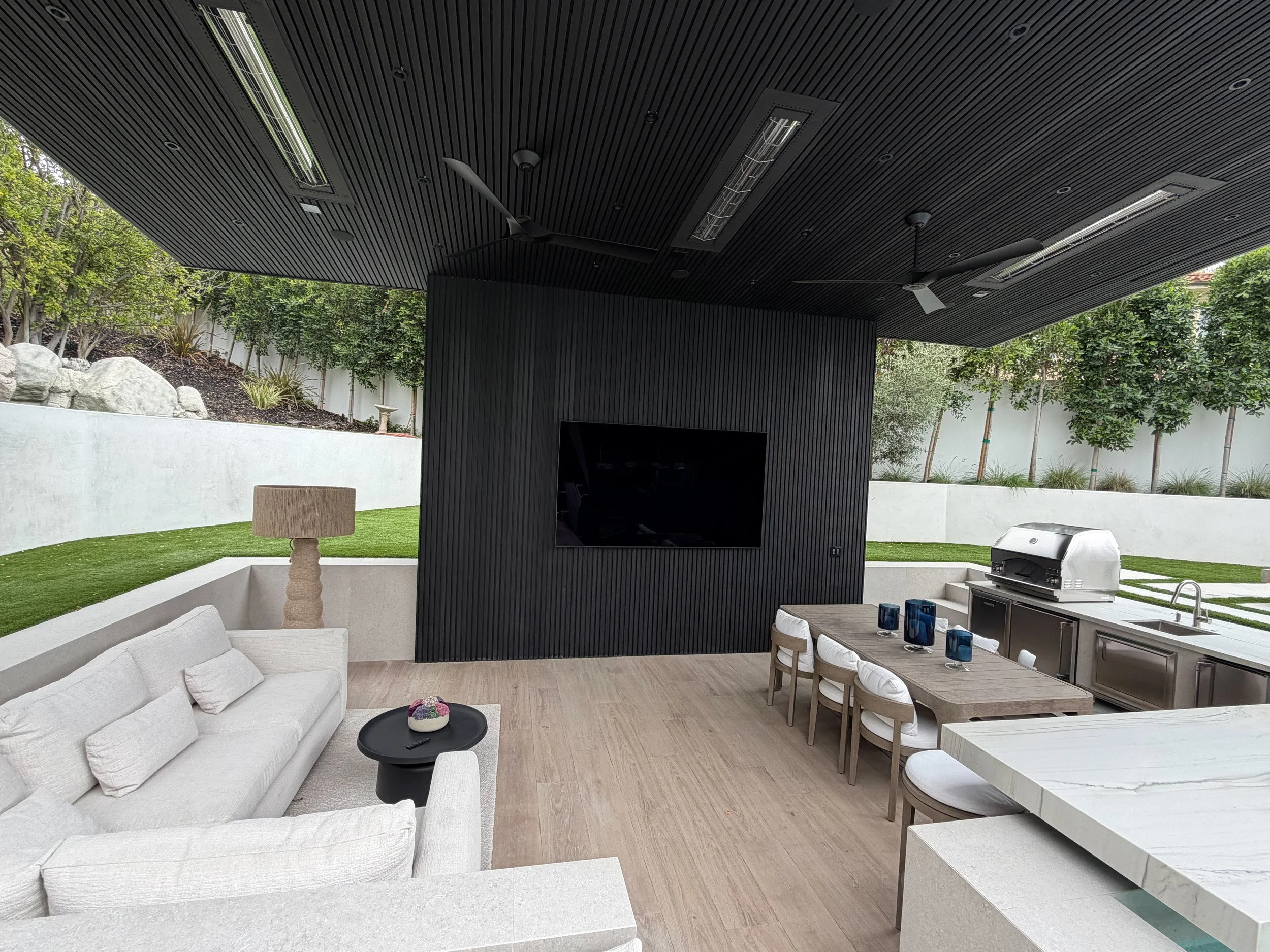 Modern outdoor patio space with white cushioned sofa, black coffee table with colorful decorative item, dining table with white chairs, built-in grill, and large black freestanding entertainment center. The space is surrounded by green trees and a wh