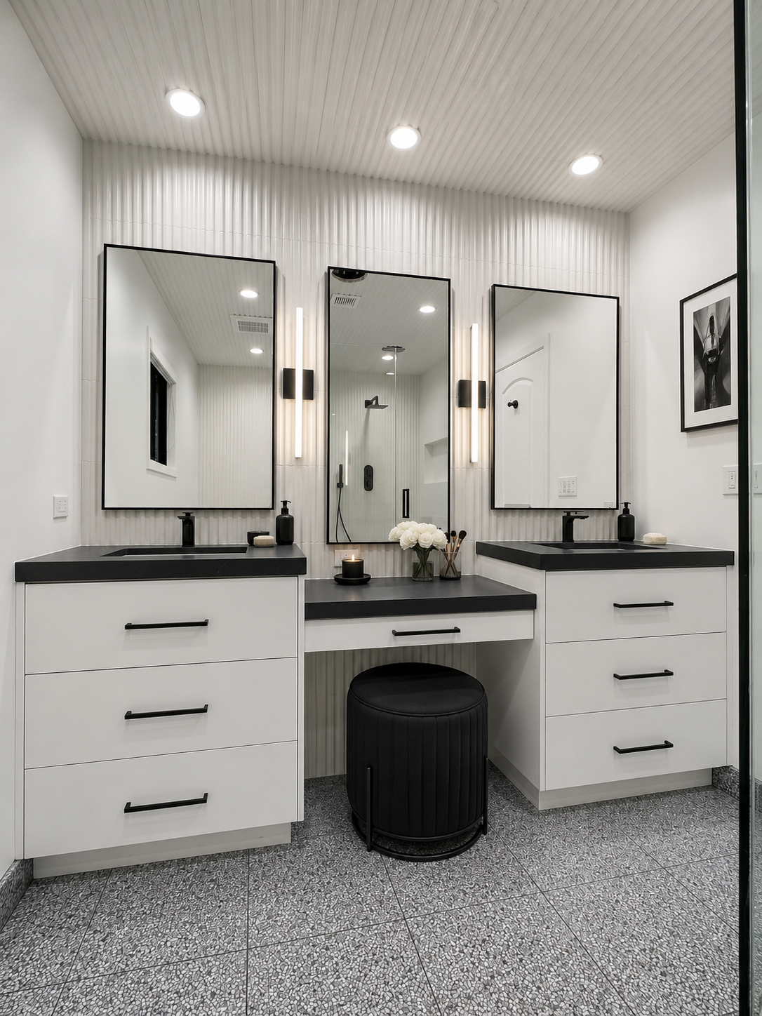 Modern black and white bathroom with two sinks, large mirrors, vertical lighting, flowers, candle, and black stool.