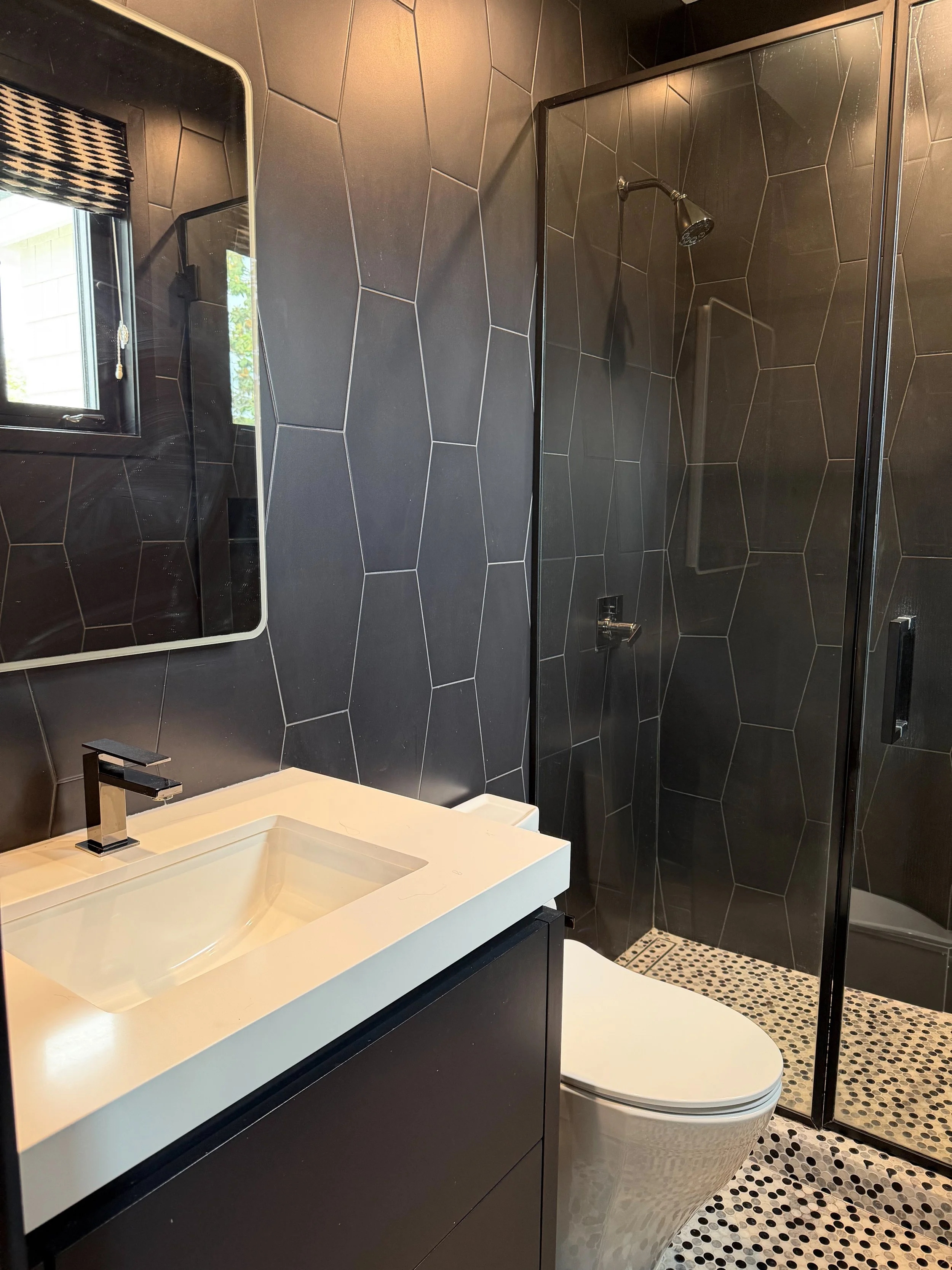 Modern bathroom with black hexagonal tile walls, a black vanity with a white integrated sink, a mirror, a frosted window with a black and white roman shade, a walk-in shower with black framing, black and white pebble floor tiles, and a white toilet.