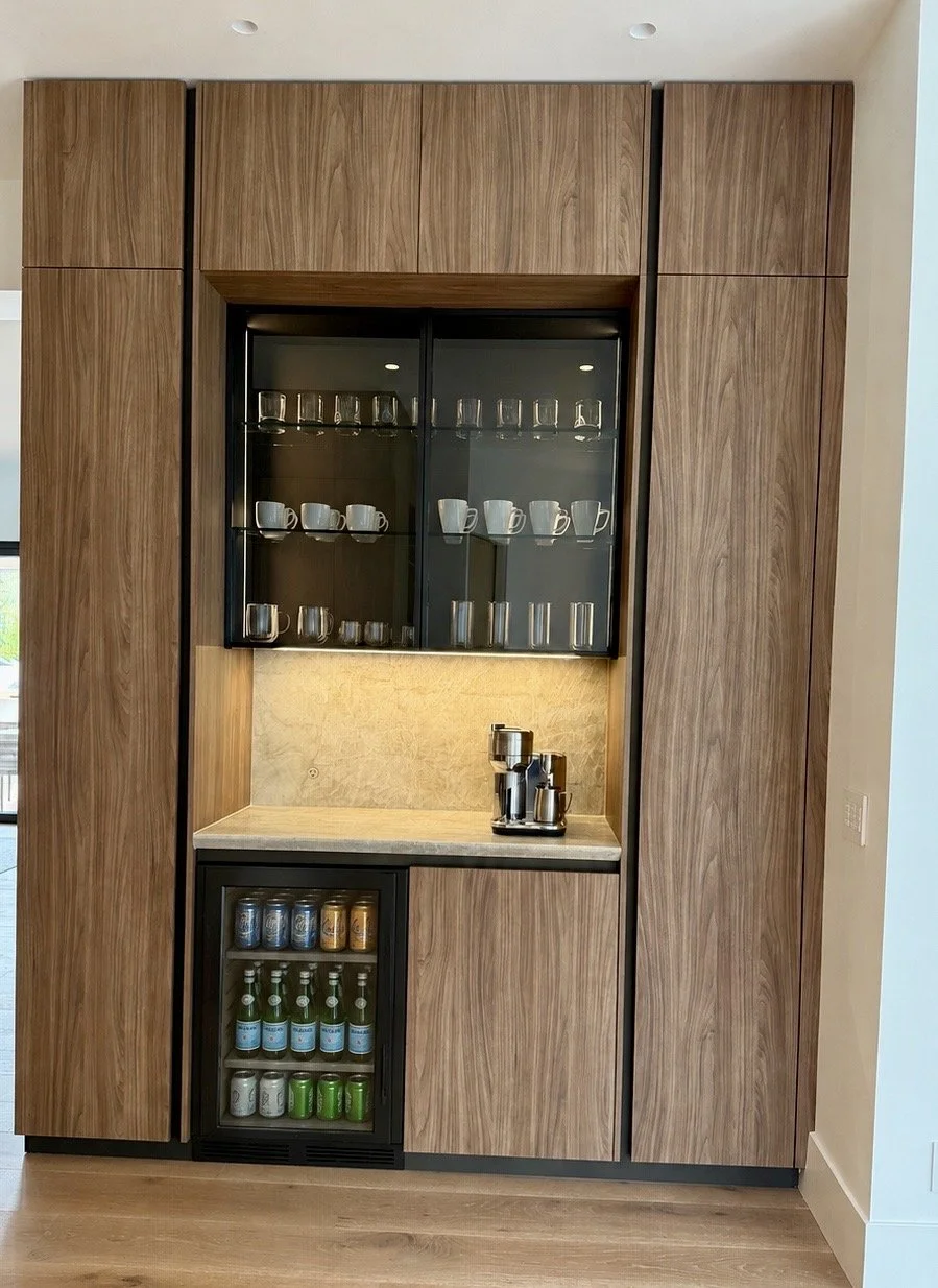 Modern wooden kitchenette with a glass-front upper cabinet holding coffee cups and glasses, a countertop with a coffee machine, and a mini fridge filled with canned and bottled drinks.