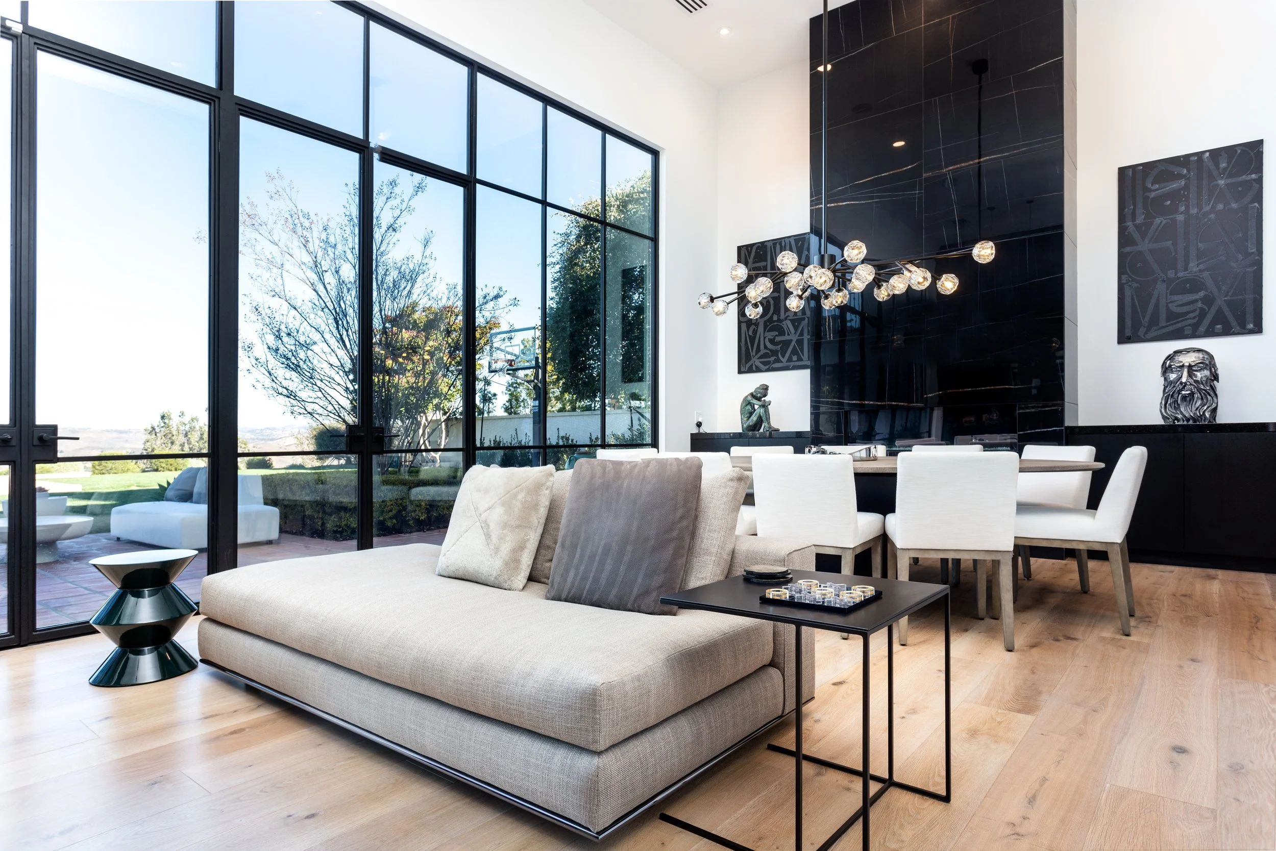 Modern living room with large floor-to-ceiling glass windows, a beige sofa with gray and beige pillows, a black side table with a geometric lamp, and a black tile accent wall with artwork and sculptures. A black chandelier hangs from the ceiling.