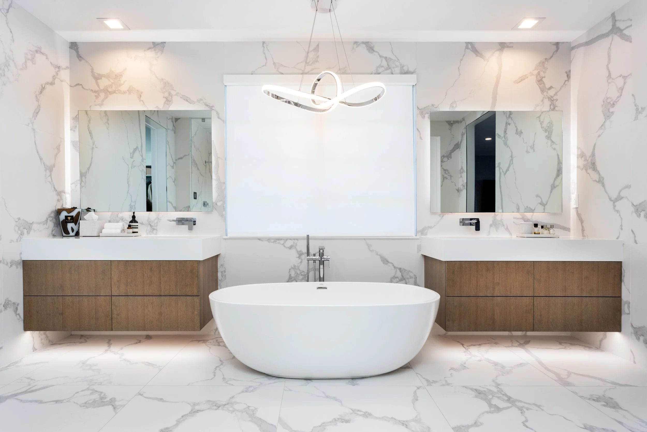 Modern bathroom with marble walls and flooring, a large white bathtub, two mirrors above dual sinks with wooden cabinets, a potted plant, and a unique futuristic chandelier hanging from the ceiling.