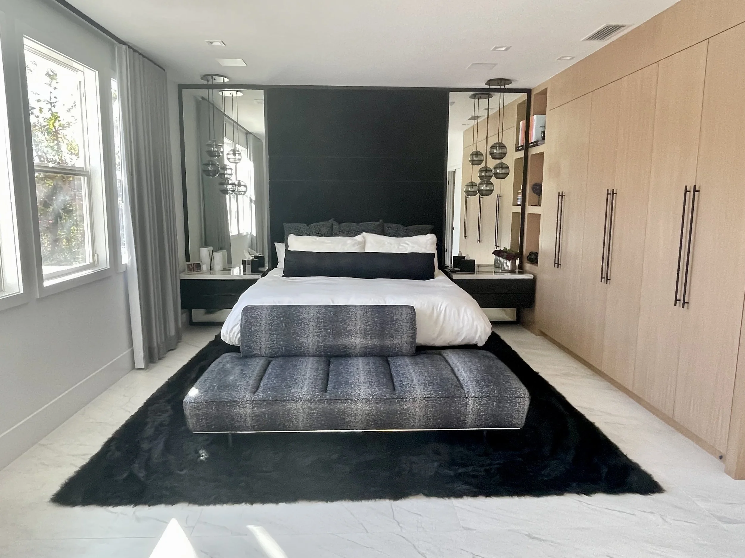 Modern bedroom with a bed featuring white bedding and dark pillows, black nightstands, large windows with white curtains, wooden built-in wardrobe, black fur rug, and black textured bench at the foot of the bed.