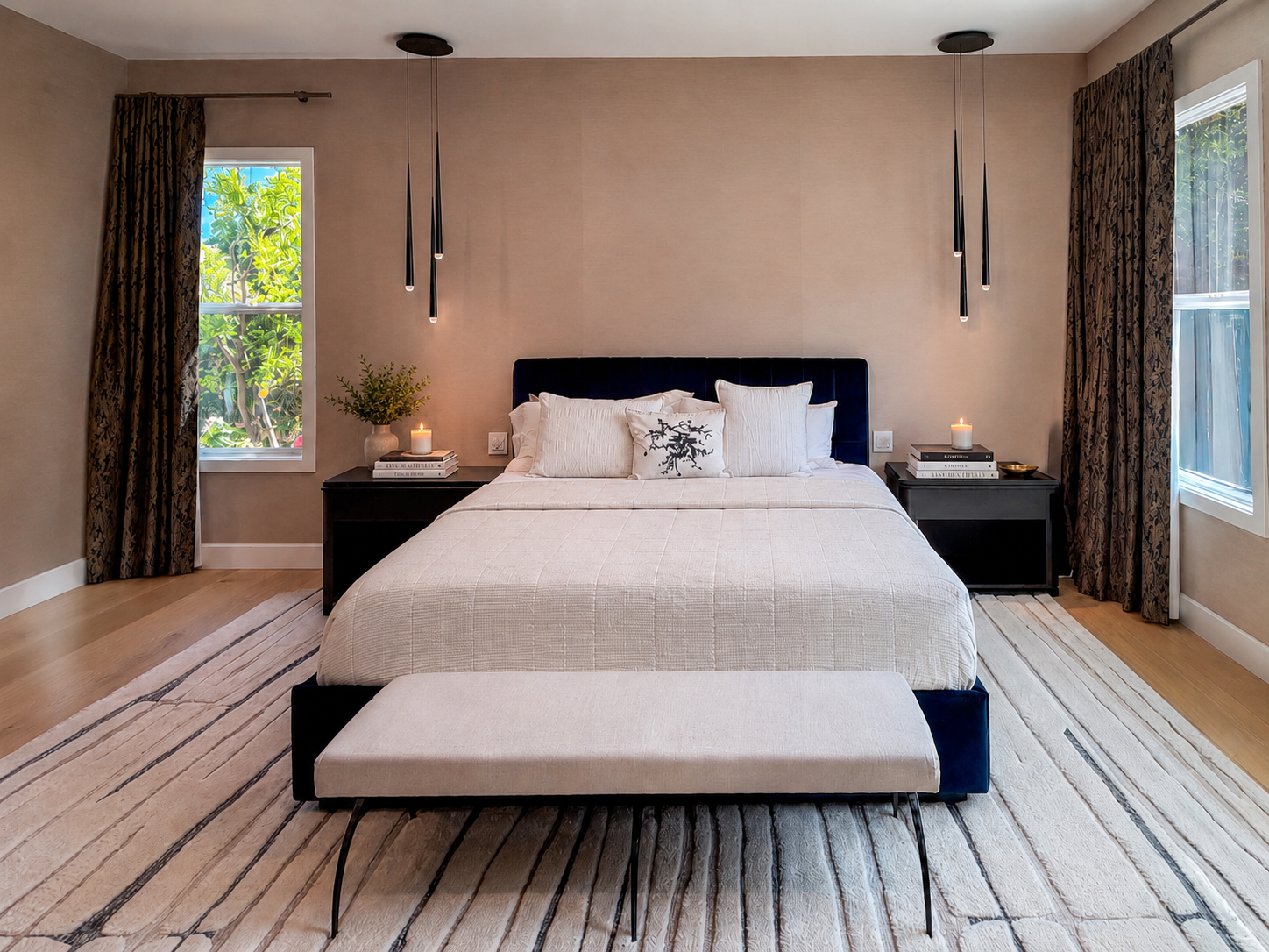 A modern bedroom with beige walls, two windows with floral curtains, a king-sized bed with a dark blue headboard, white bedding, and decorative pillows. There are two black nightstands on either side of the bed, each with a candle and books. A beige 