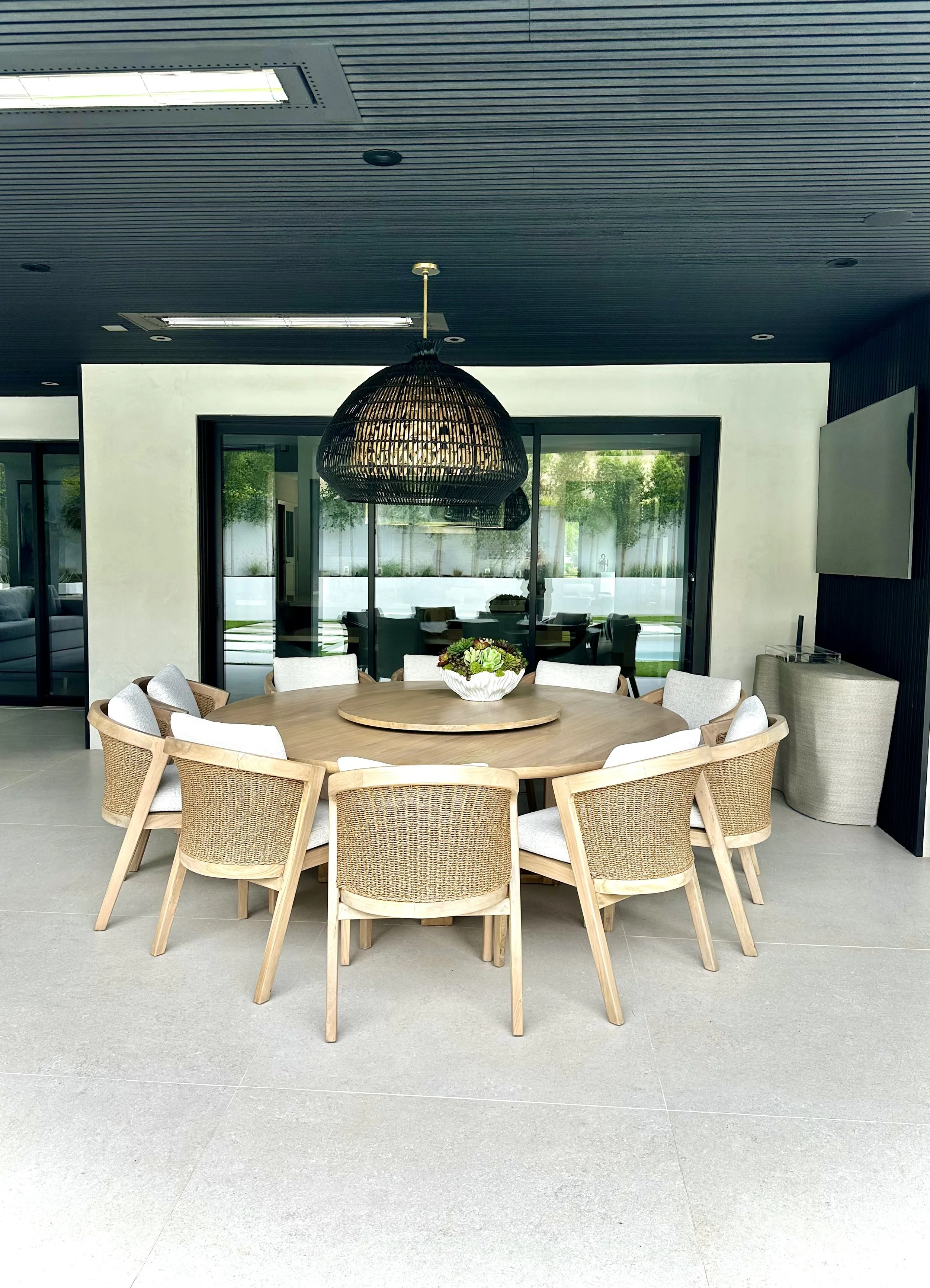 Indoor dining area featuring a large round wooden table surrounded by wicker and upholstered chairs, with a decorative centerpiece, large glass sliding doors in the background, a black pendant light over the table, and a dark wooden ceiling with buil