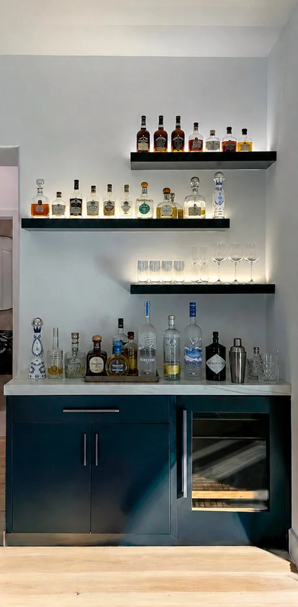 A home bar with three black shelves holding various bottles of liquor, empty wine glasses, and cocktail glasses. The bar has a blue cabinet with silver handles and a built-in mini fridge below.