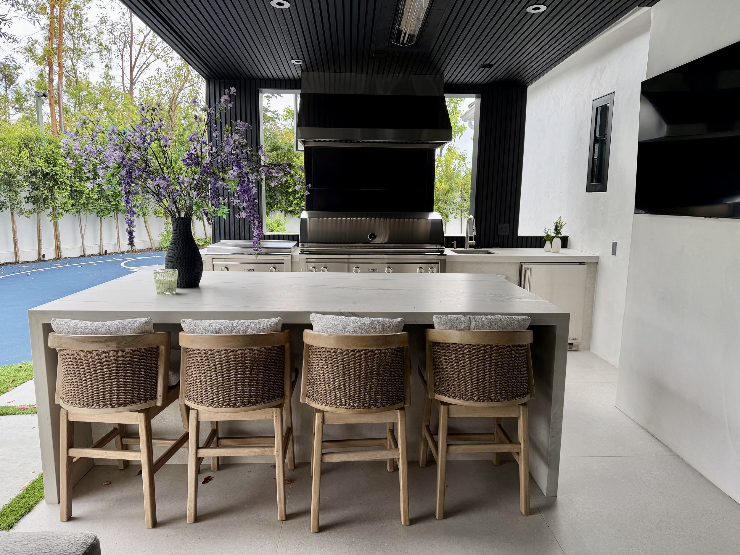 A modern outdoor kitchen with a large white dining table, six wicker chairs, a black vase with purple flowers, stainless steel grill, black vent hood, black wood paneling, white walls, and a flat-screen TV, with trees visible outside.