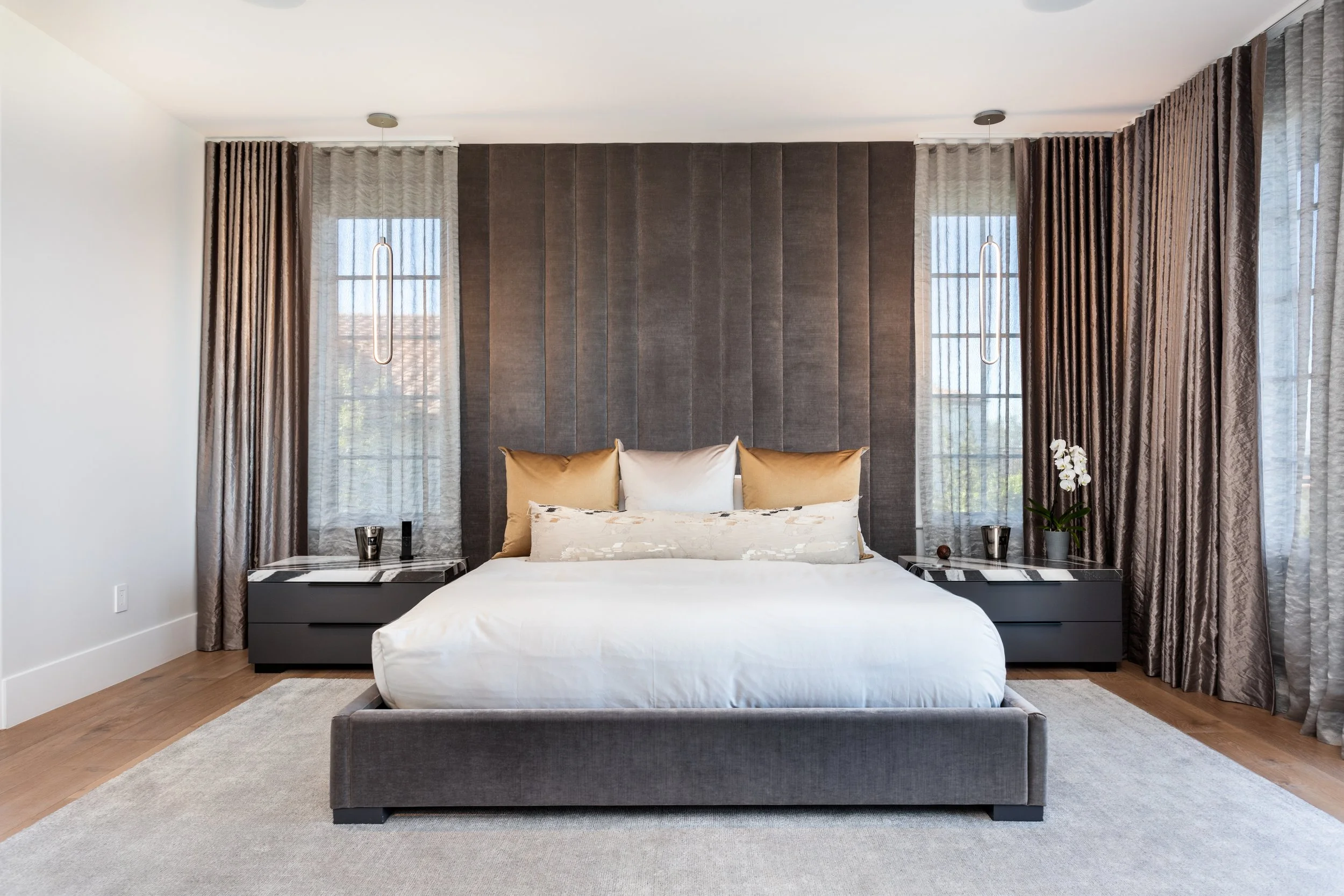 Modern bedroom with a gray upholstered bed, beige and white pillows, dark gray nightstands, and tall windows with sheer curtains and brown drapes, illuminated by pendant lights.