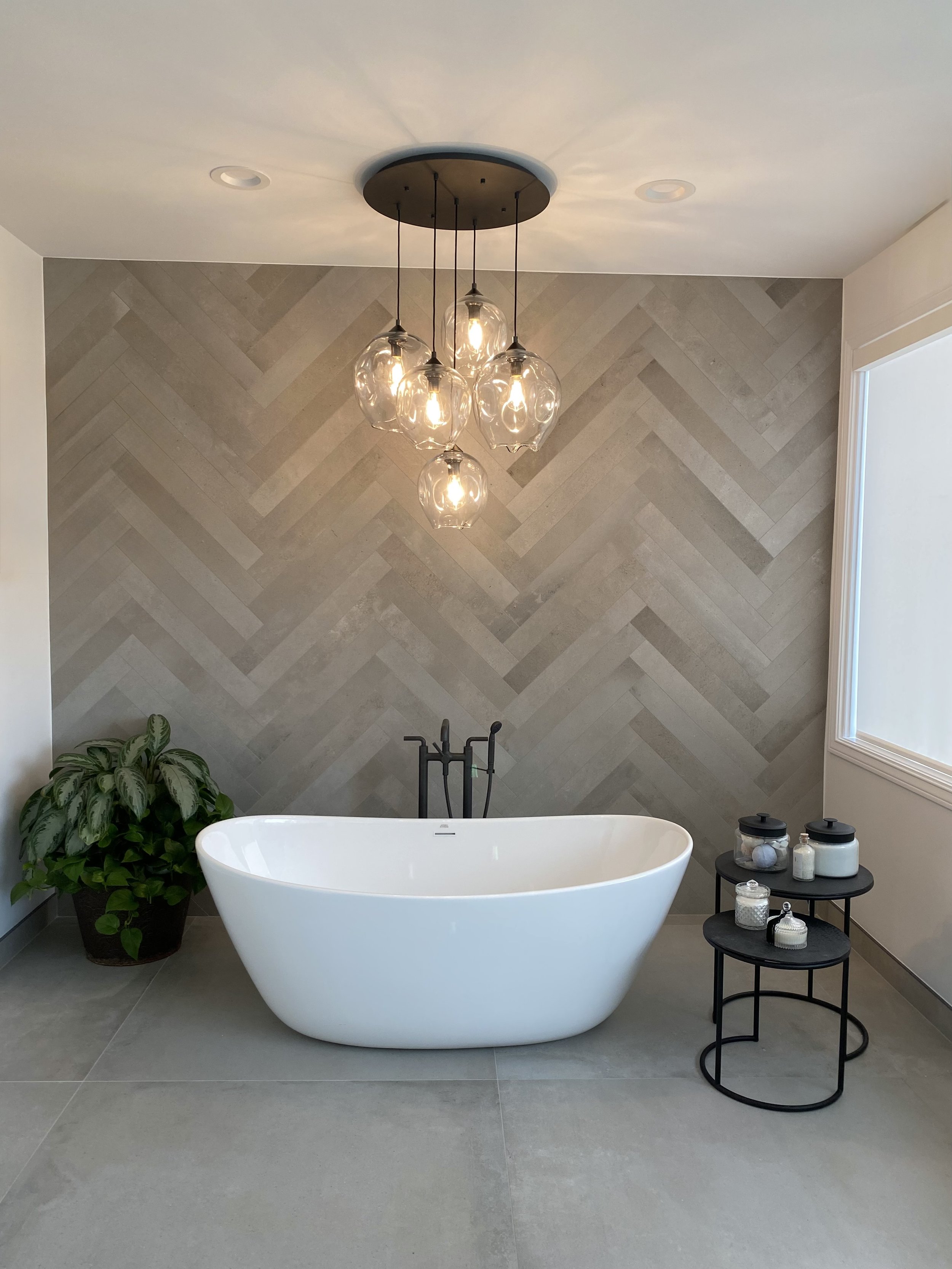 Modern bathroom with a white freestanding bathtub, a green potted plant, a black side table with jars and bottles, a geometric tile wall, a window, and a ceiling light fixture with multiple glass globes.