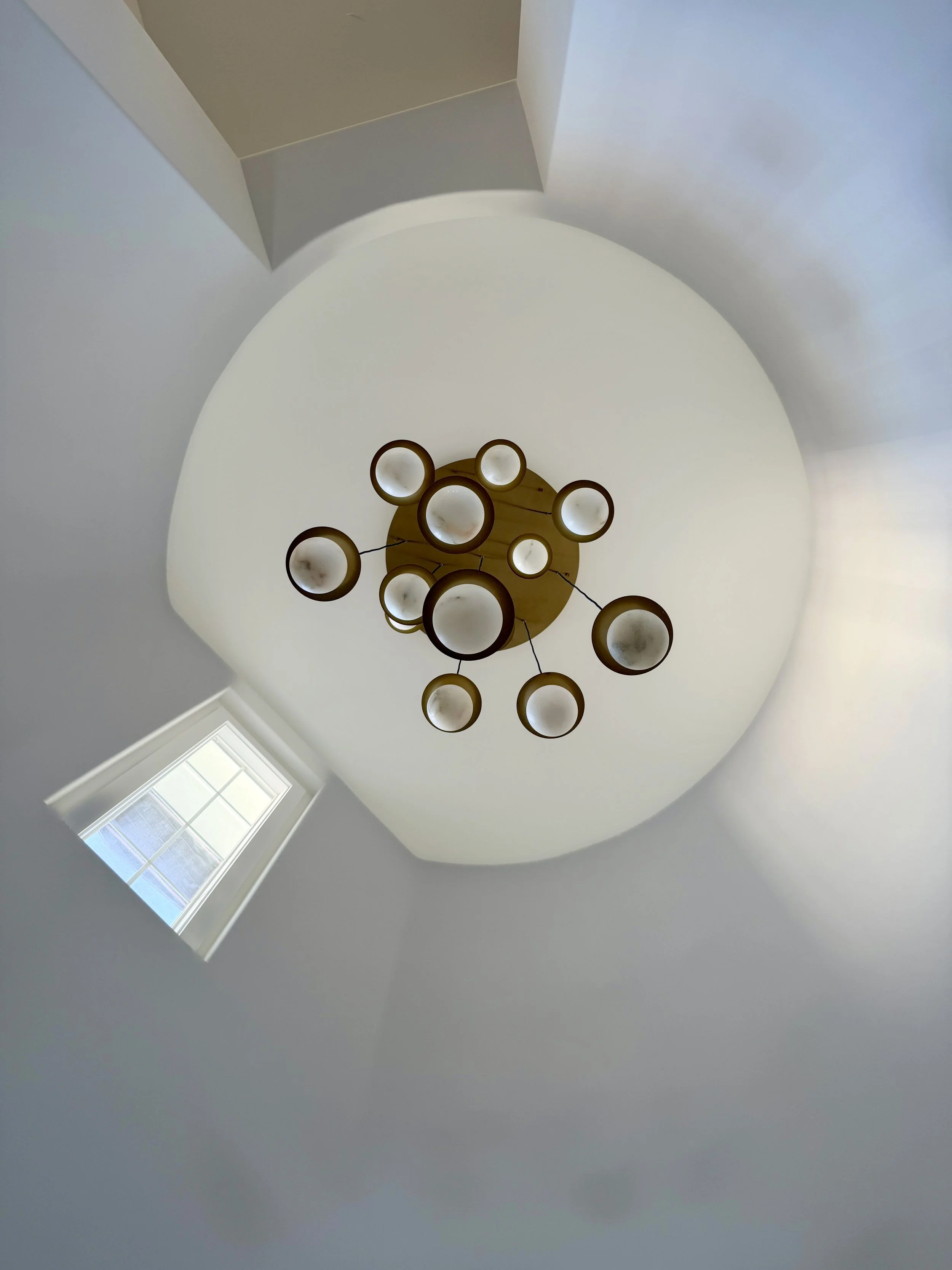 Looking up at a circular hanging chandelier with multiple lights in a house with a window.