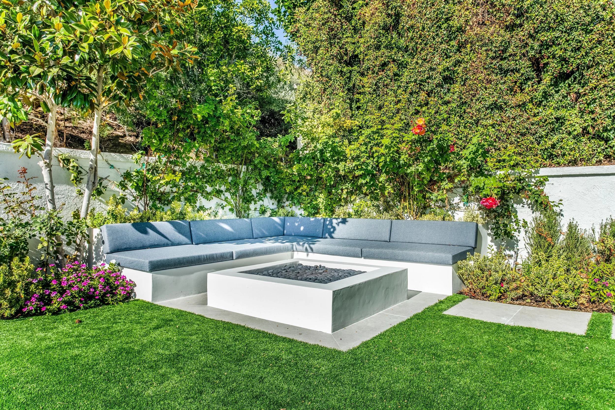 A modern outdoor seating area with a curved gray cushioned bench surrounding a white firepit, set in a lush green garden with various plants, shrubs, and a hedge background.