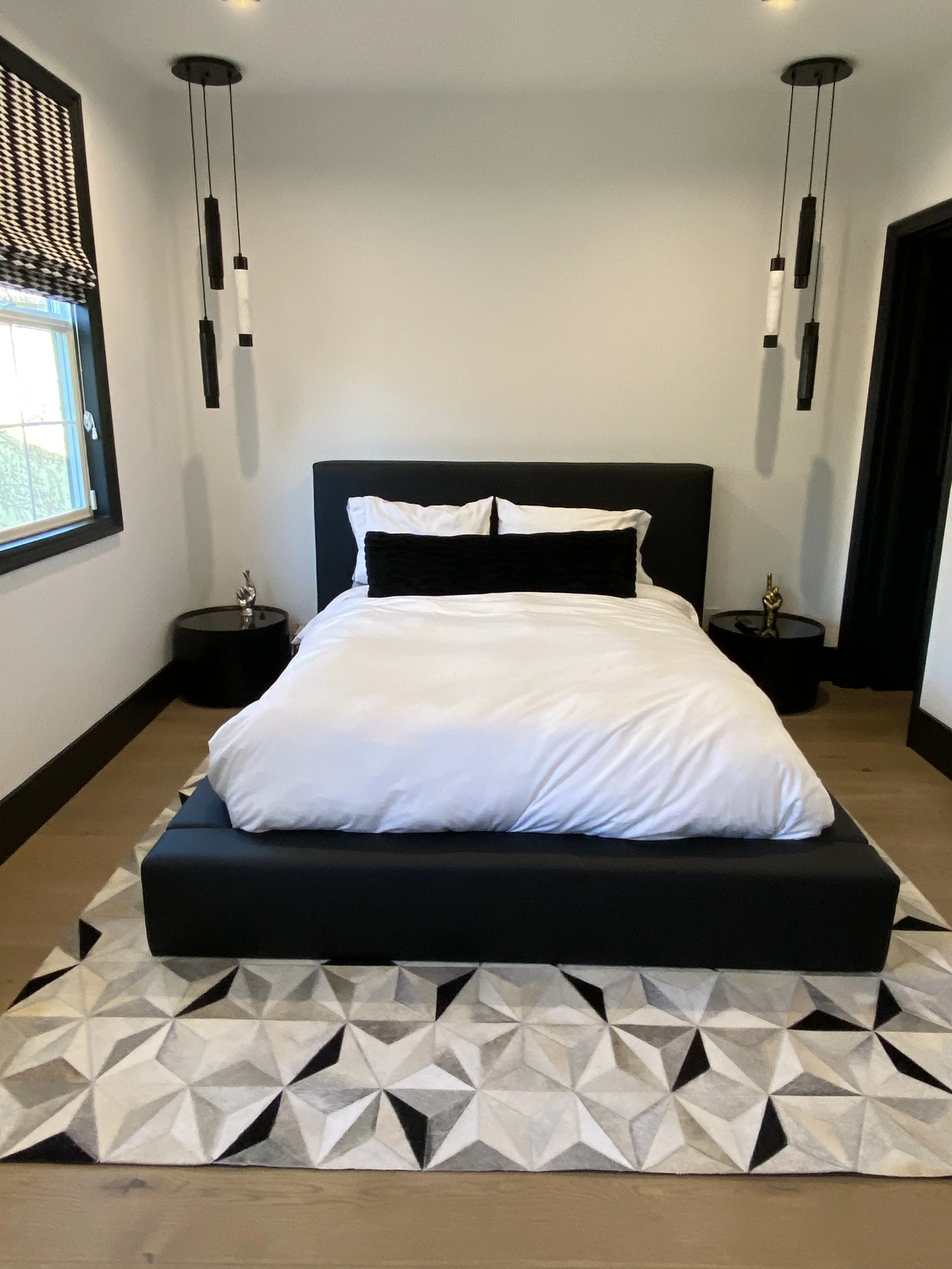A bedroom with a bed, black headboard, white bedding, and black pillow. Two black nightstands with decorative items, black pendant lights hanging from the ceiling, a window with a patterned black-and-white Roman shade, and a geometric patterned rug o