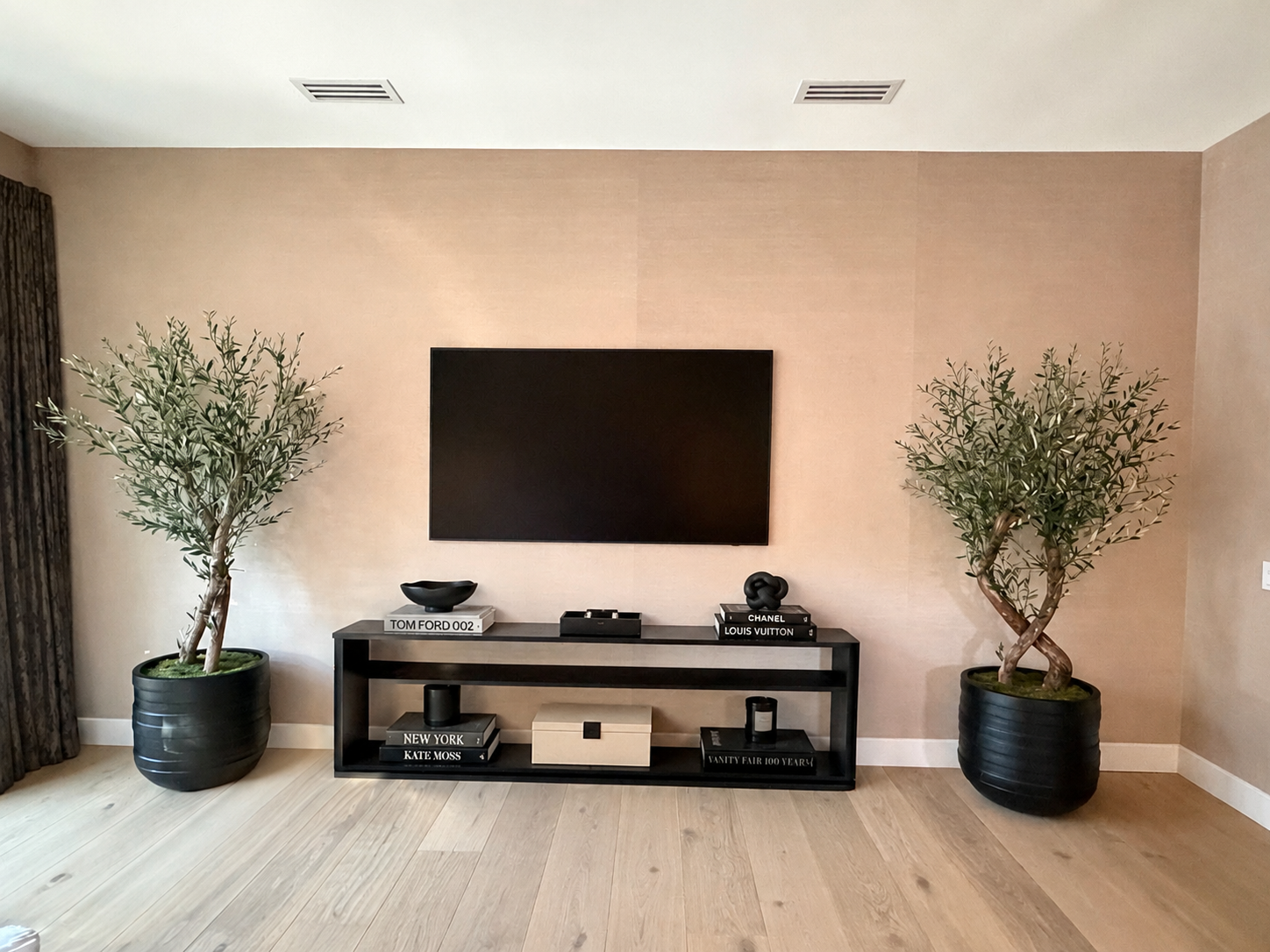 Interior living room with beige walls, a black TV on the wall, a black console table with decorative books, a bowl, and sculpture, flanked by two large potted olive trees, and dark curtains on the left.