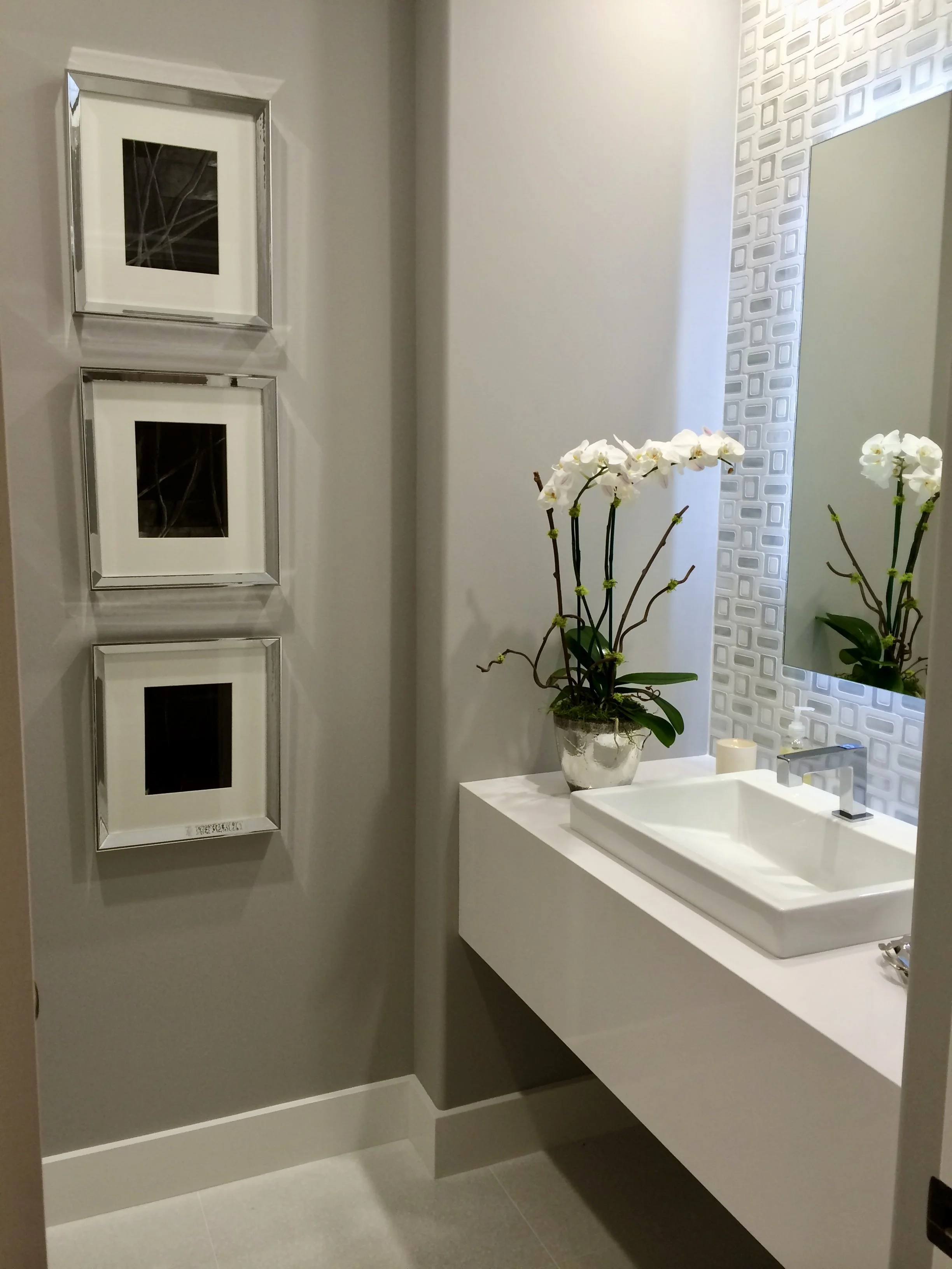A modern bathroom with a white vanity sink, a mirror with a textured white frame, white orchids in a glass vase, and two black-and-white framed pictures on a gray wall.