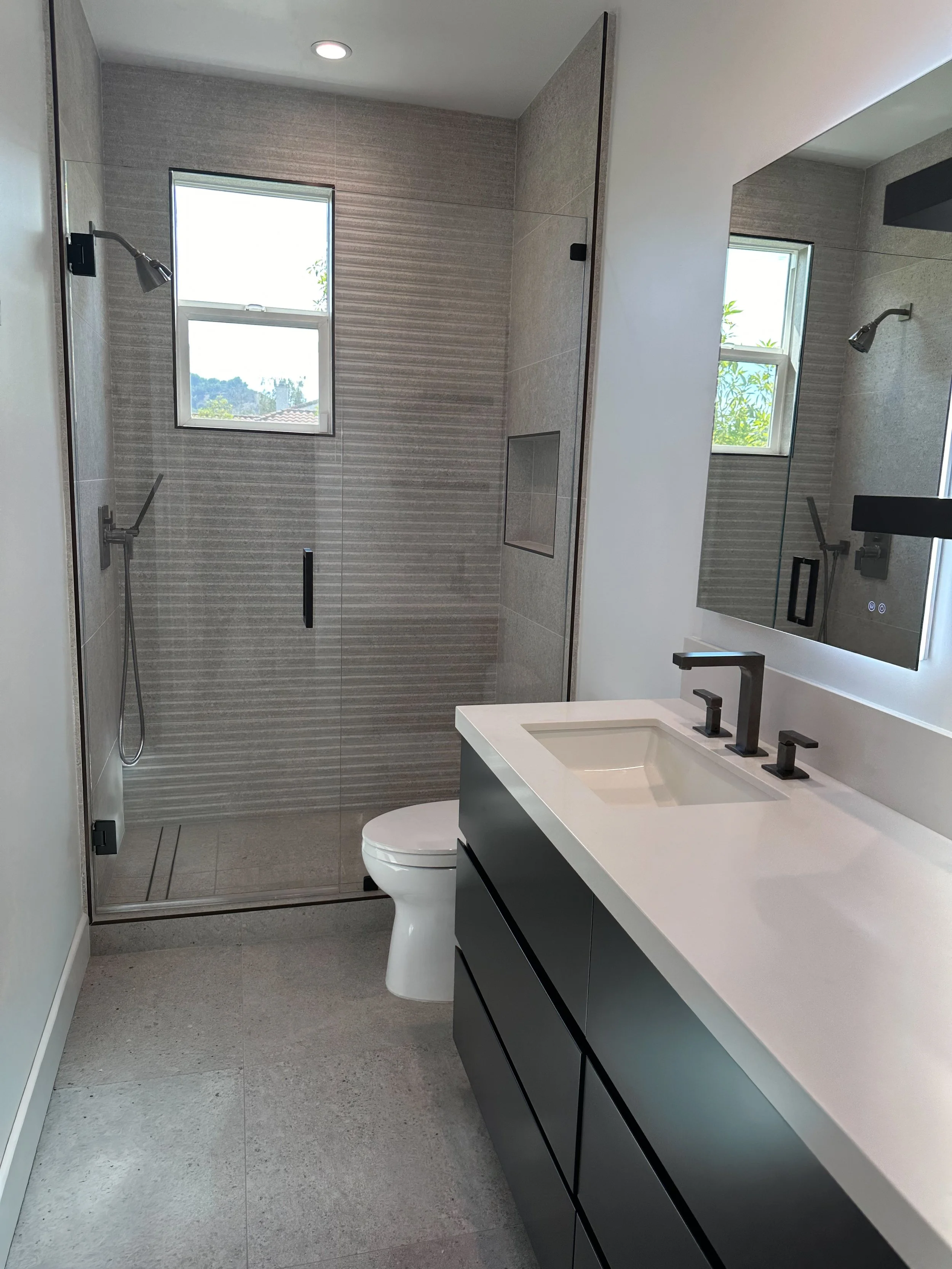 Modern bathroom with a walk-in shower, a white vanity with a black cabinet, and a toilet. The shower has glass doors, two small windows with greenery outside, and neutral-toned tile walls. A large mirror is above the sink.