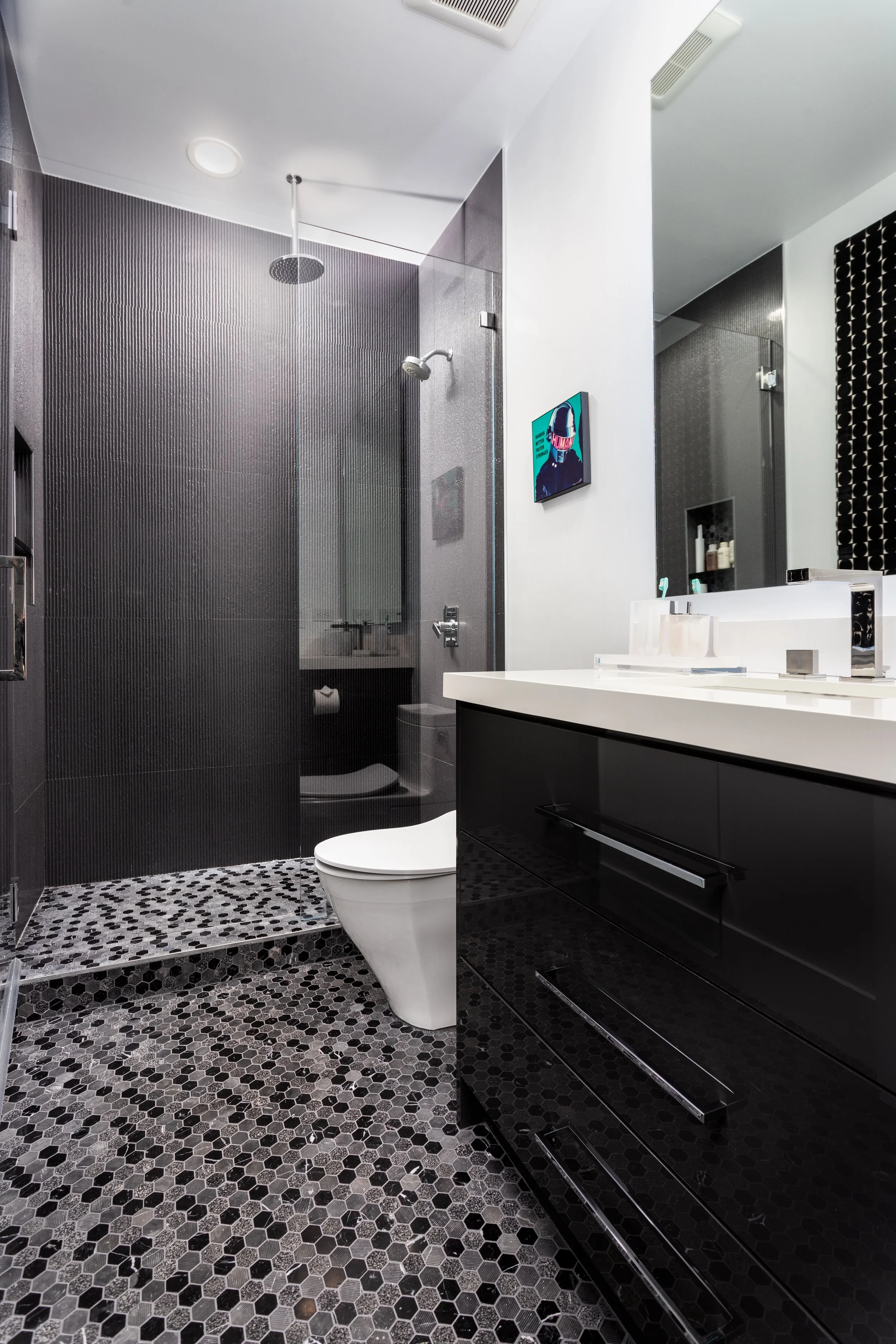 Modern bathroom with black and white hexagonal patterned floor tiles, black vanity with drawers, white countertop with two sinks, wall-mounted faucets, large mirror, shower area with dark textured wall tiles, and a colorful artwork on the wall.