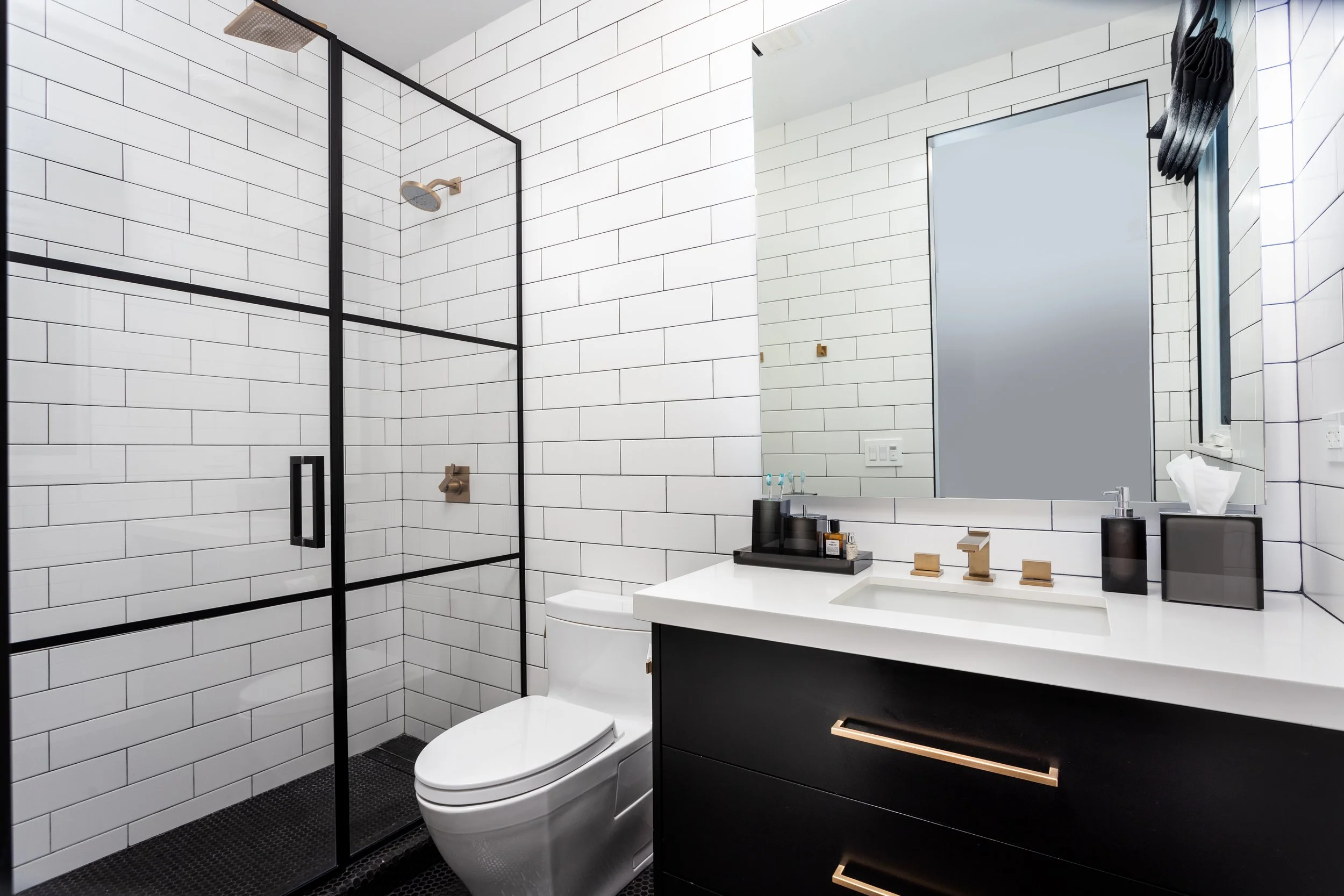 Modern bathroom with white subway tile walls, a glass-enclosed shower, a wall-mounted faucet, a large mirror, a black vanity with gold accents, and a white toilet.