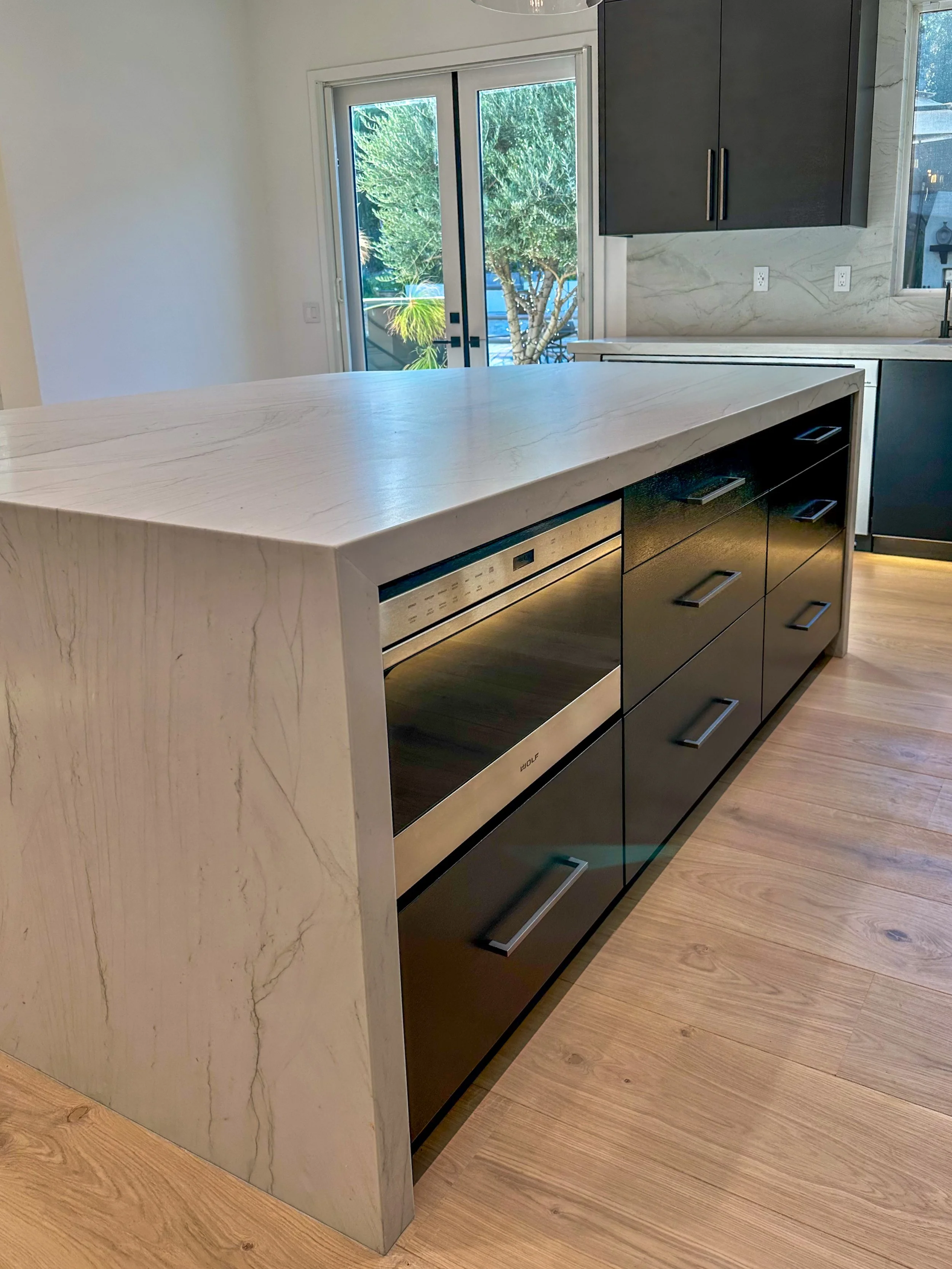 Modern kitchen with a white marble countertop island, black cabinets, a built-in microwave, and sliding glass doors leading to an outdoor space with trees.