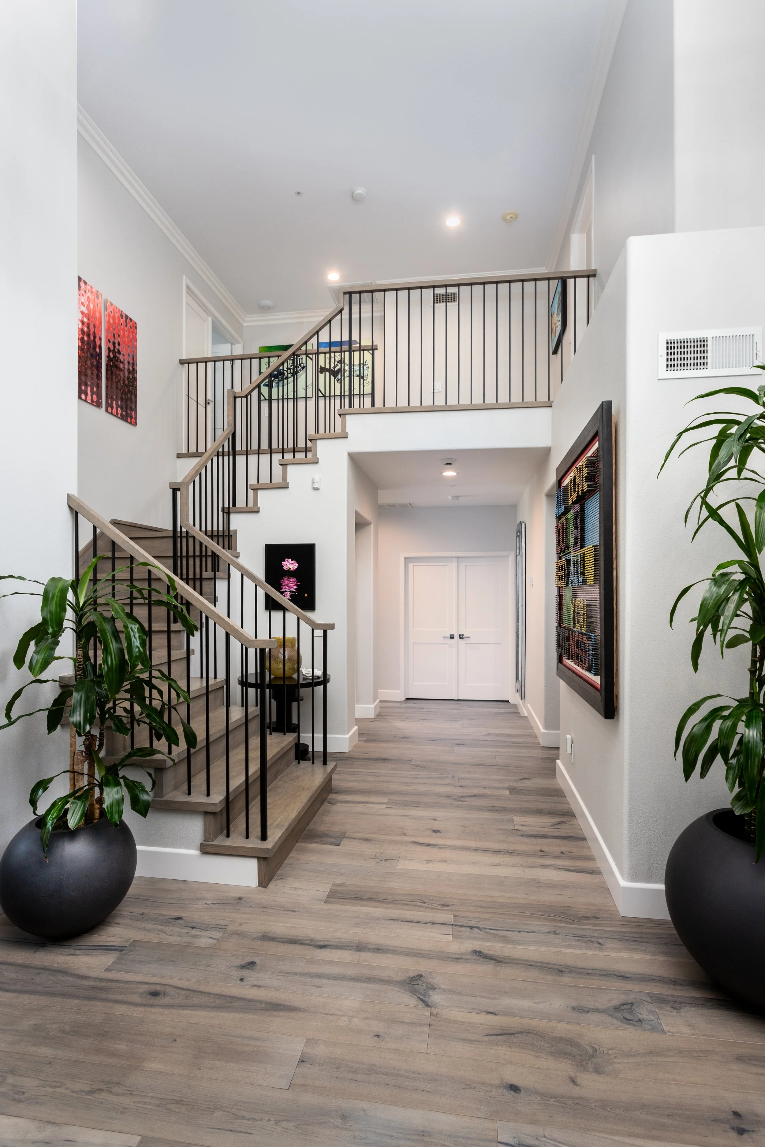 Interior take of a modern house entryway with stairs, black railing, wooden floors, and artwork on the white walls with potted plants.
