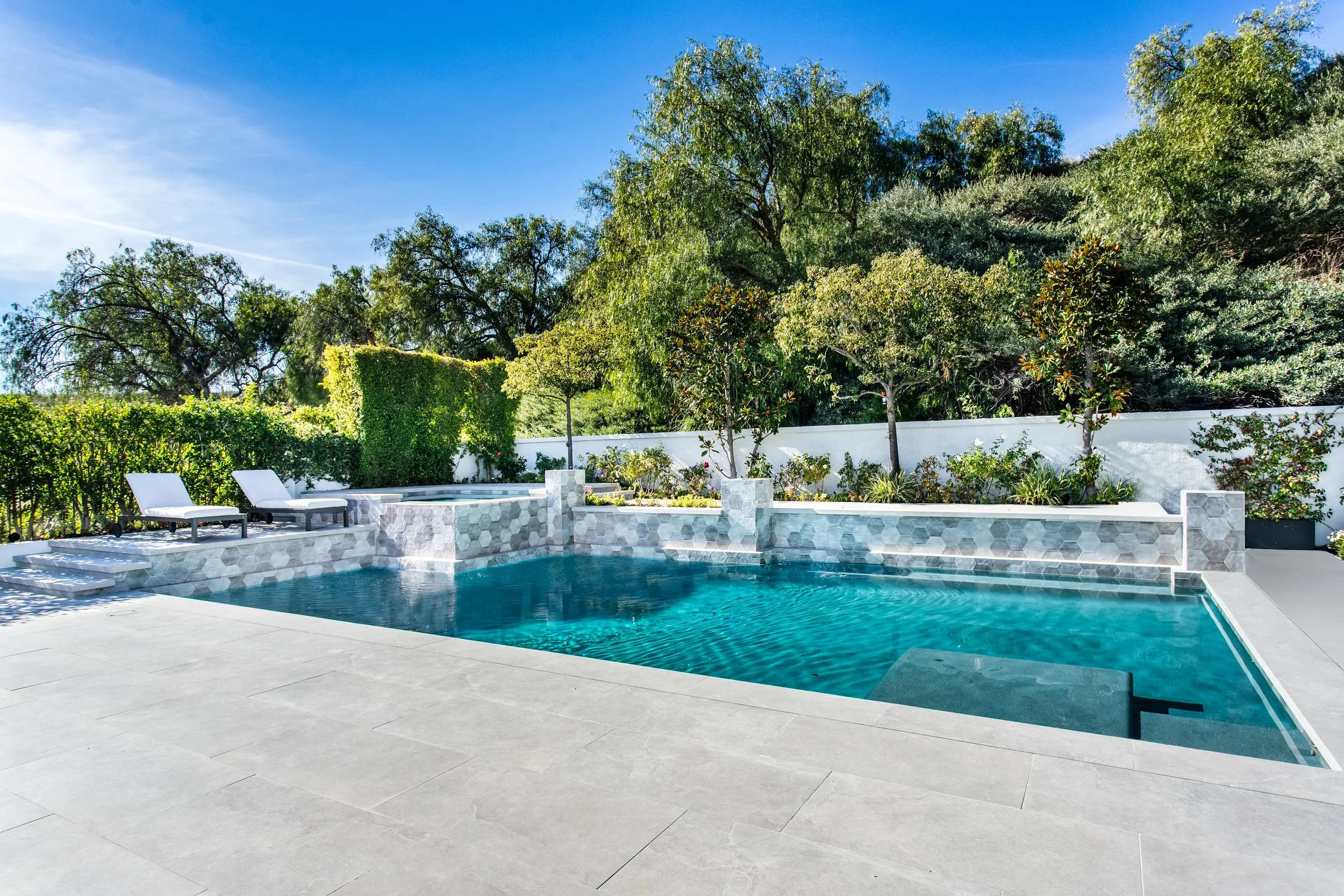 A backyard with a swimming pool, surrounded by white concrete, with lounge chairs on the deck and lush green trees in the background under a blue sky.