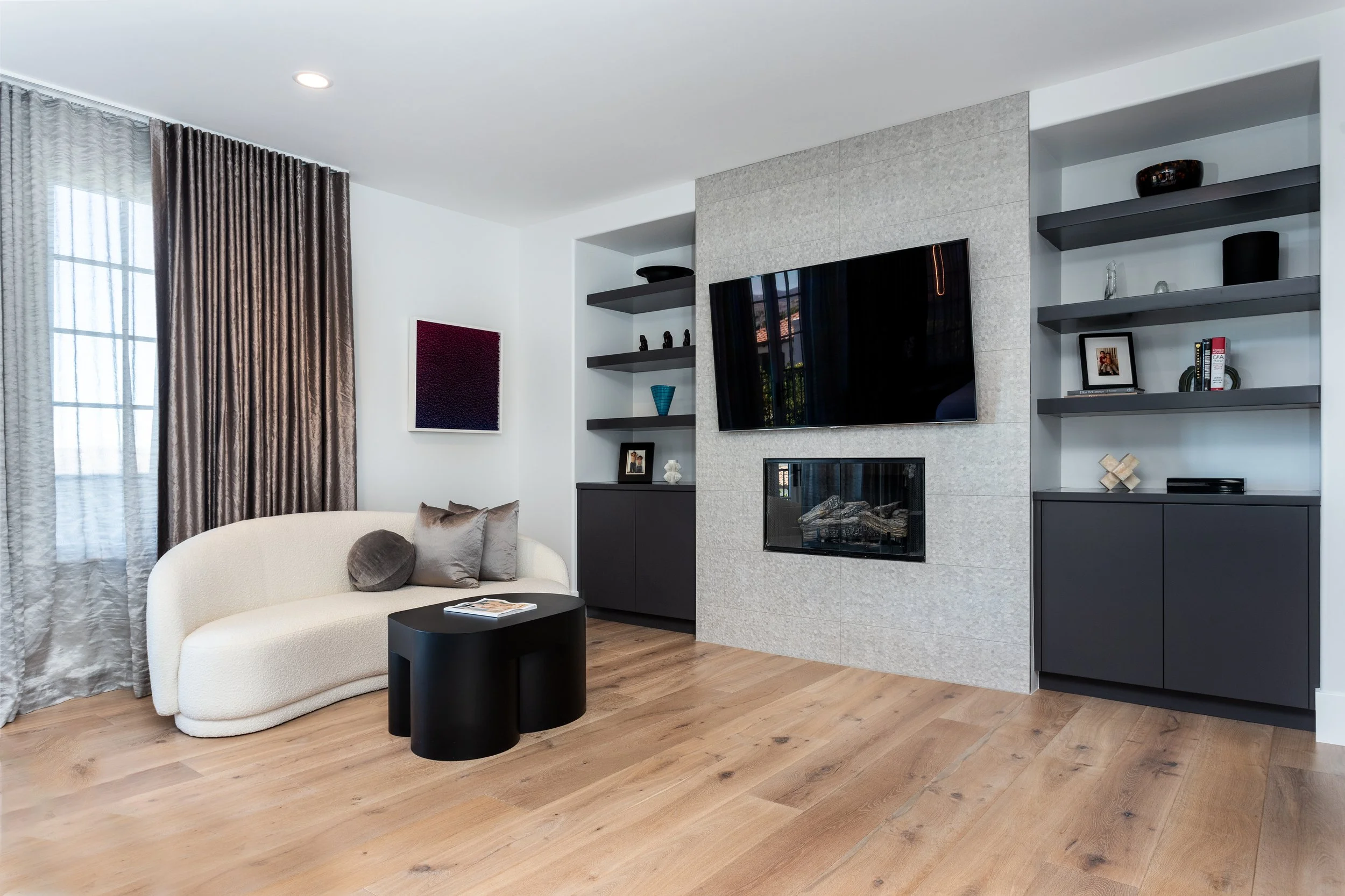 Living room with white sofa, black coffee table, built-in shelves with decor, large window with two sets of curtains, television on textured wall, hardwood floors.
