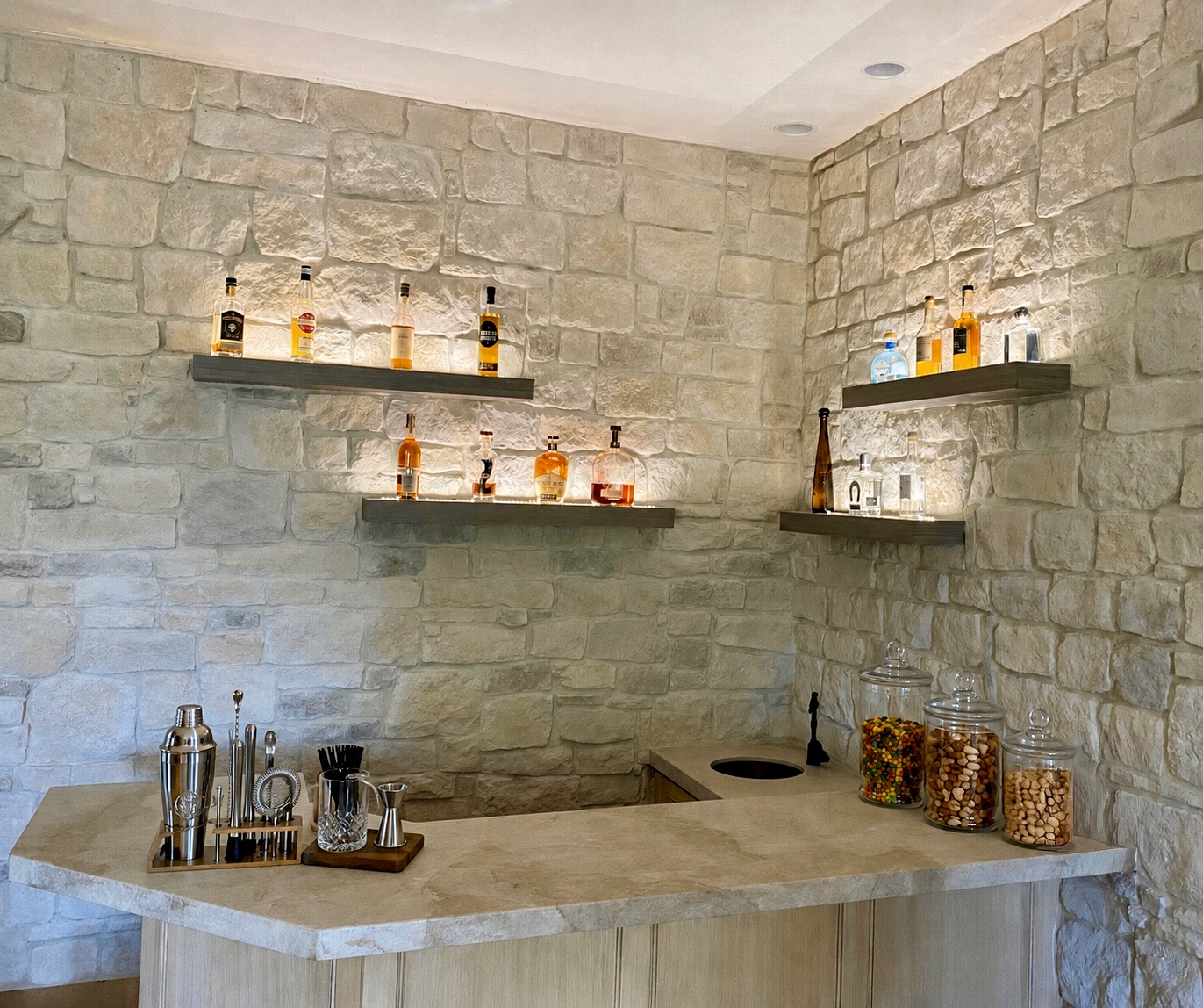 A bar area with a stone wall background, shelves holding various bottles of alcohol, jars of candies, and a marble countertop with bar tools.