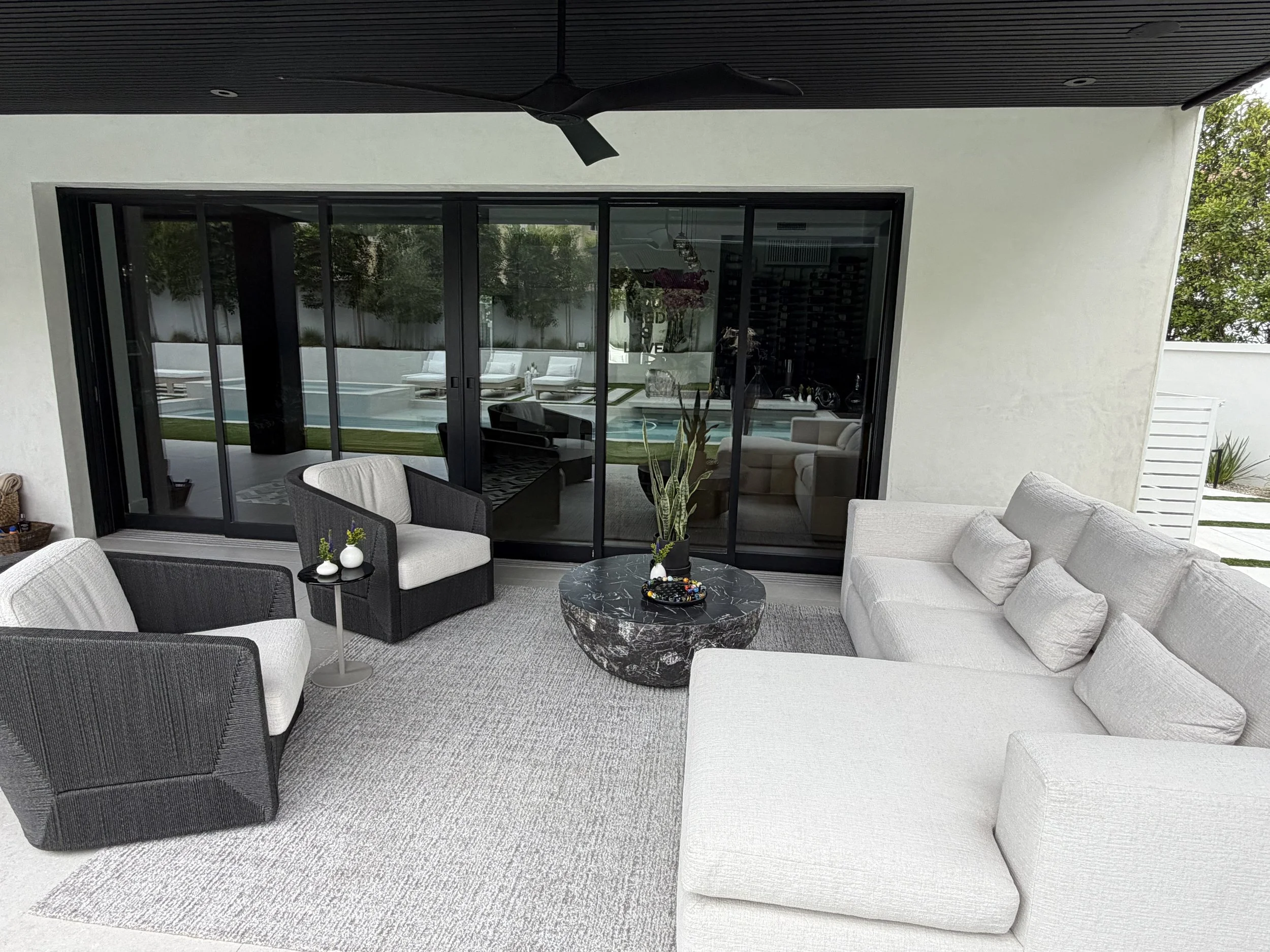 A spacious outdoor patio with white and black seating, a black marble coffee table, and a view of a backyard with a pool and lounge chairs through large glass sliding doors.