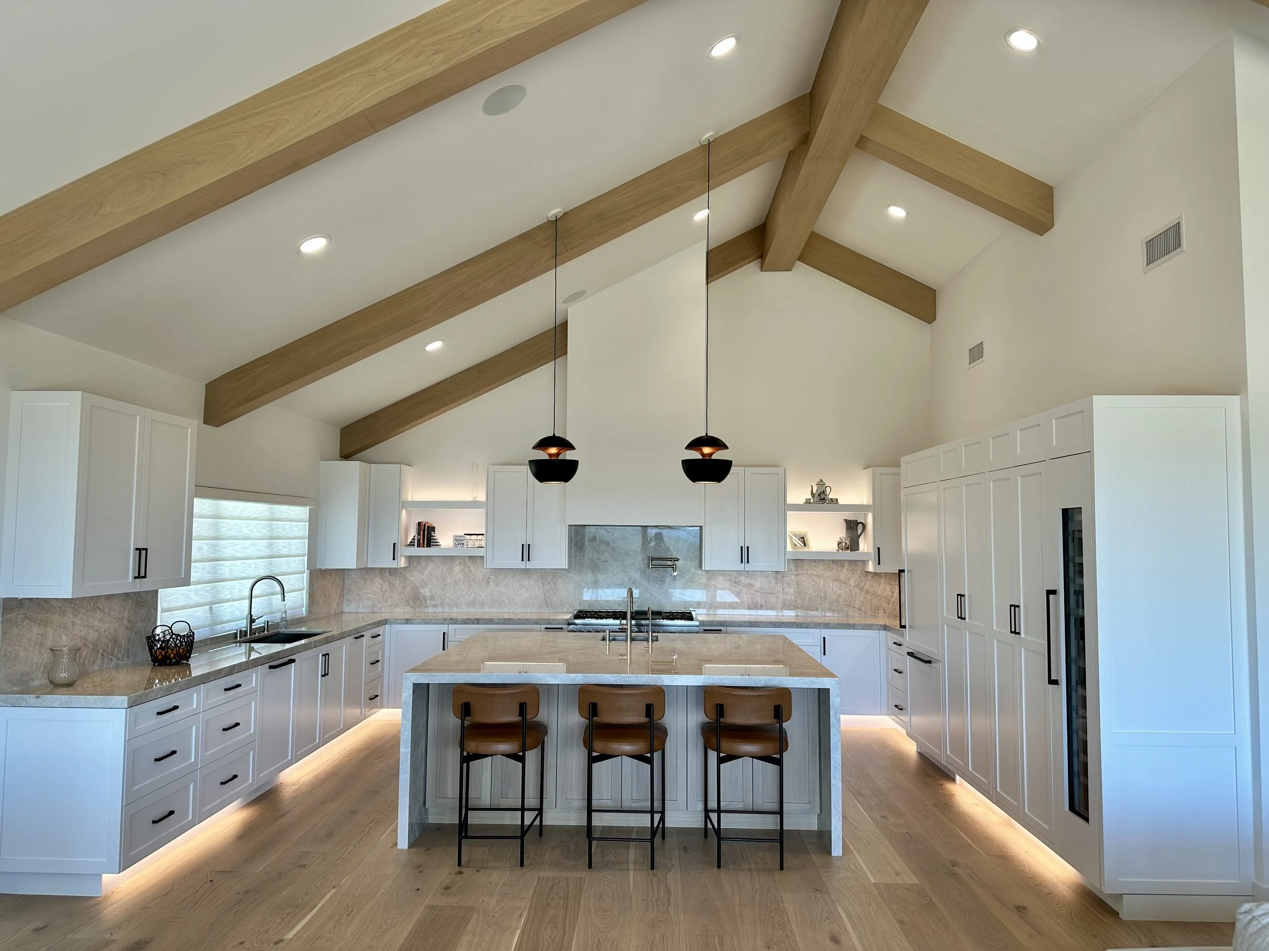 Modern kitchen with white cabinets, a large marble island with seating, pendant lights, and an open vaulted ceiling with wooden beams.