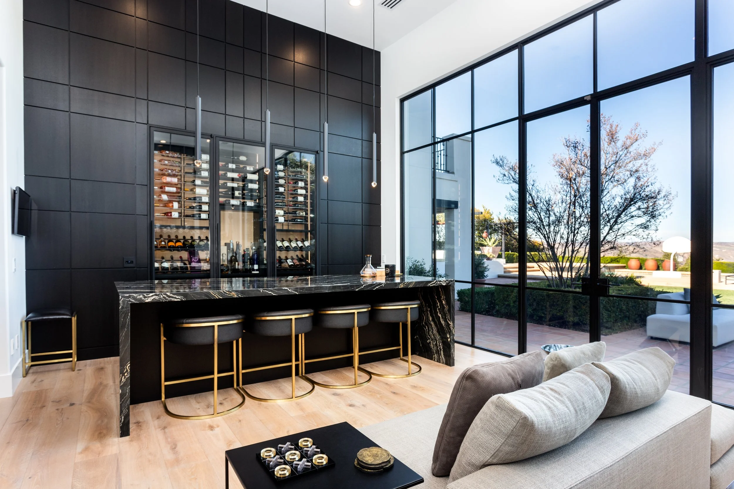 Modern living room with a black wall featuring a built-in wine refrigerator, a marble kitchen island with four black and gold barstools, and large floor-to-ceiling windows showing an outdoor patio with trees and planters.