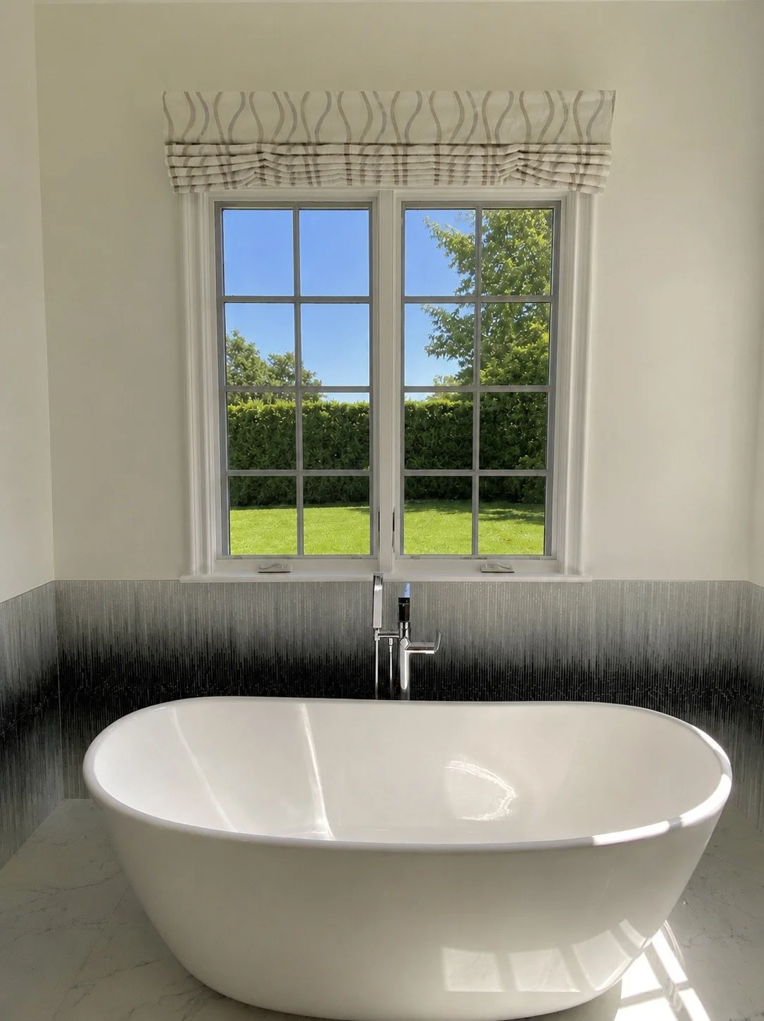 A modern bathroom with a freestanding white bathtub, large double-paned window with striped window valance, allowing natural light, and a view of a green lawn and trees outside.