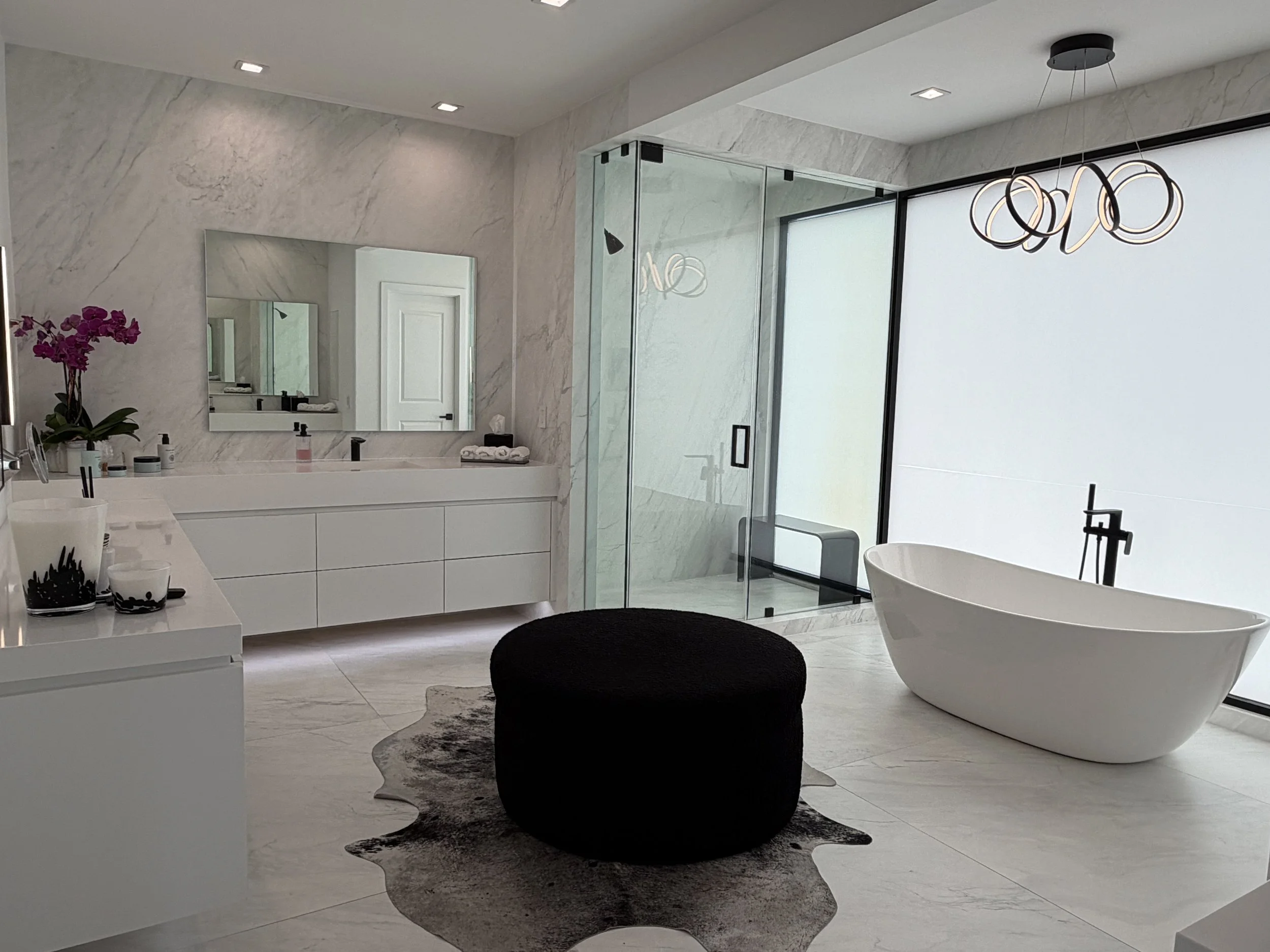 Modern bathroom featuring a white bathtub, cowhide rug, black ottoman, large mirror, purple orchid plant, glass shower enclosure, and contemporary light fixture.