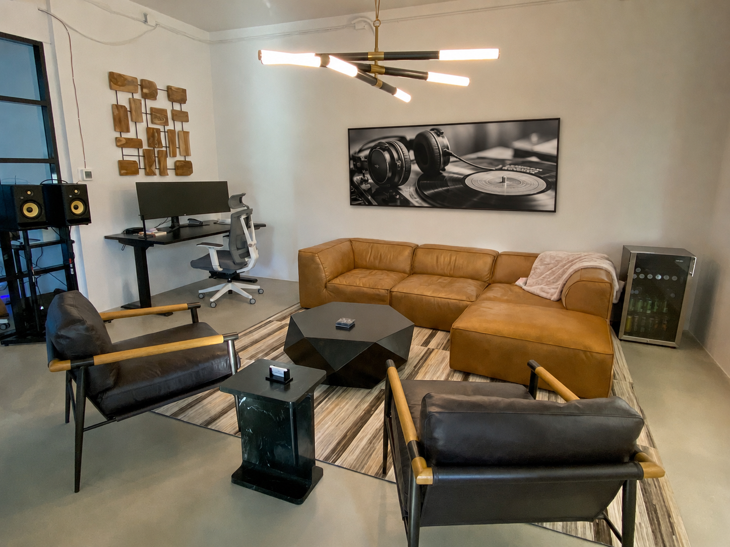 Modern living room with a brown leather sectional sofa, black armchairs with wooden arms, a geometric black coffee table, and a patterned area rug. A framed black and white photo of a turntable and headphones hangs on the wall. A desk with two large 