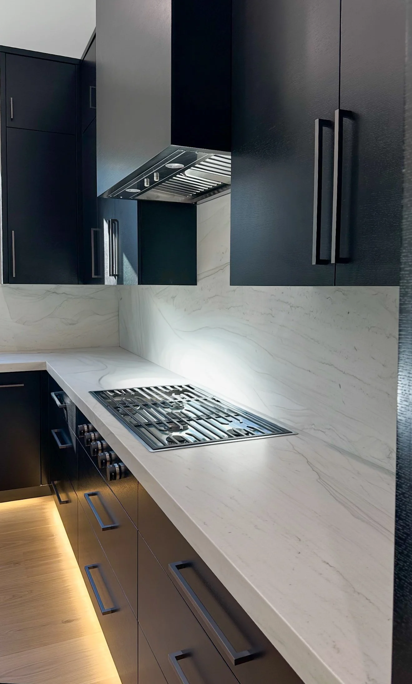 A modern kitchen with black cabinetry, a white marble countertop with gray veining, a built-in stainless steel gas cooktop, and a white marble backsplash. The cabinets have sleek, black handles, and beneath the counter, there are drawers with black h