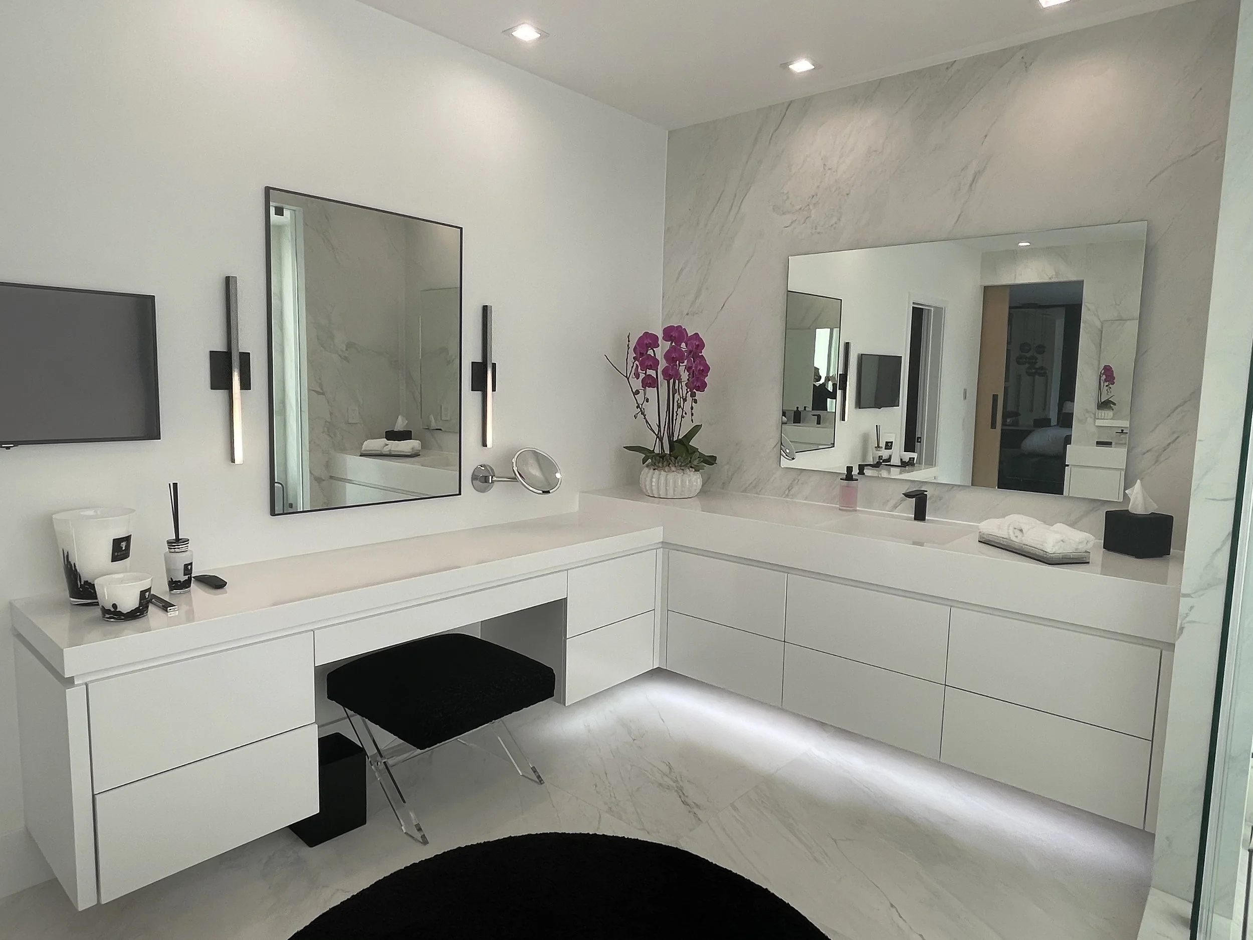 A modern bathroom vanity area with a white countertop, a large mirror, and a orchid plant in a white pot. The wall has a marble finish and there are two vertical light fixtures next to the mirror. The corner of a TV and a black stool are also visible
