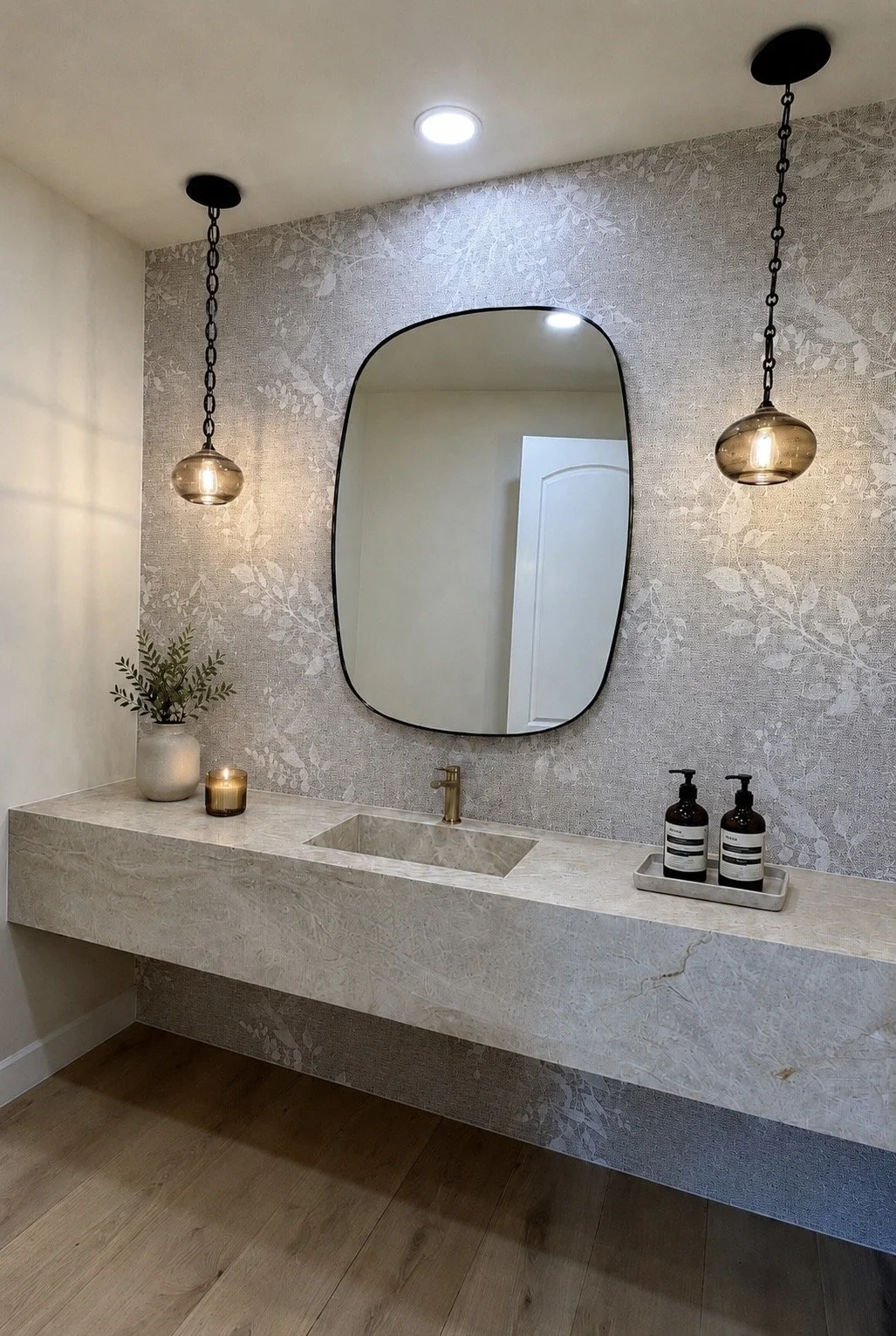 Modern bathroom vanity with a marble countertop, a gold faucet, a large oval mirror, two pendant lights, a small vase with greenery, a candle, and soap bottles on a tray.